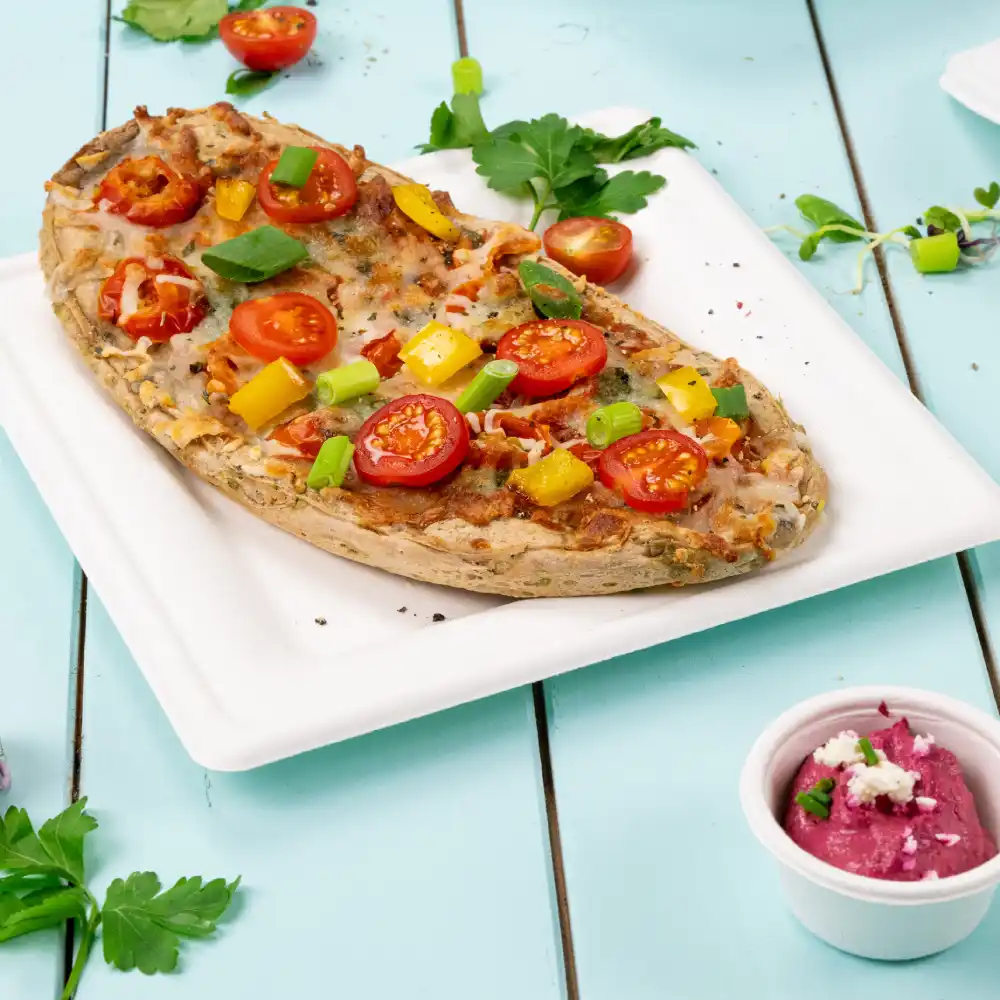 Pane piatto cotto guarnito con pomodorini, cipollotti e peperoni, servito su un piatto bianco con un contorno di salsa.