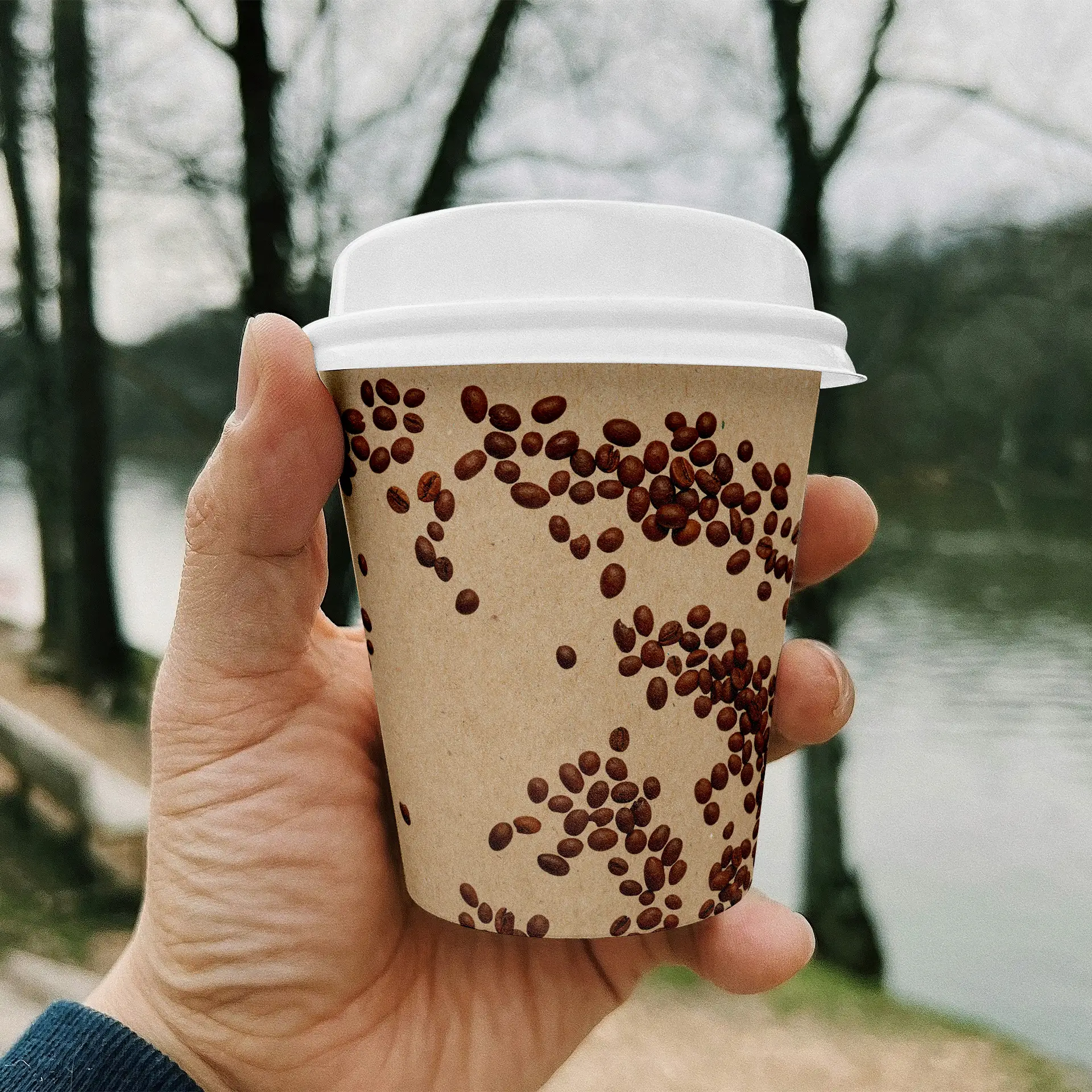 Un cup in carta marrone stampato con un design di chicchi di caffè, tenuto da una mano, su uno sfondo naturale sfocato.