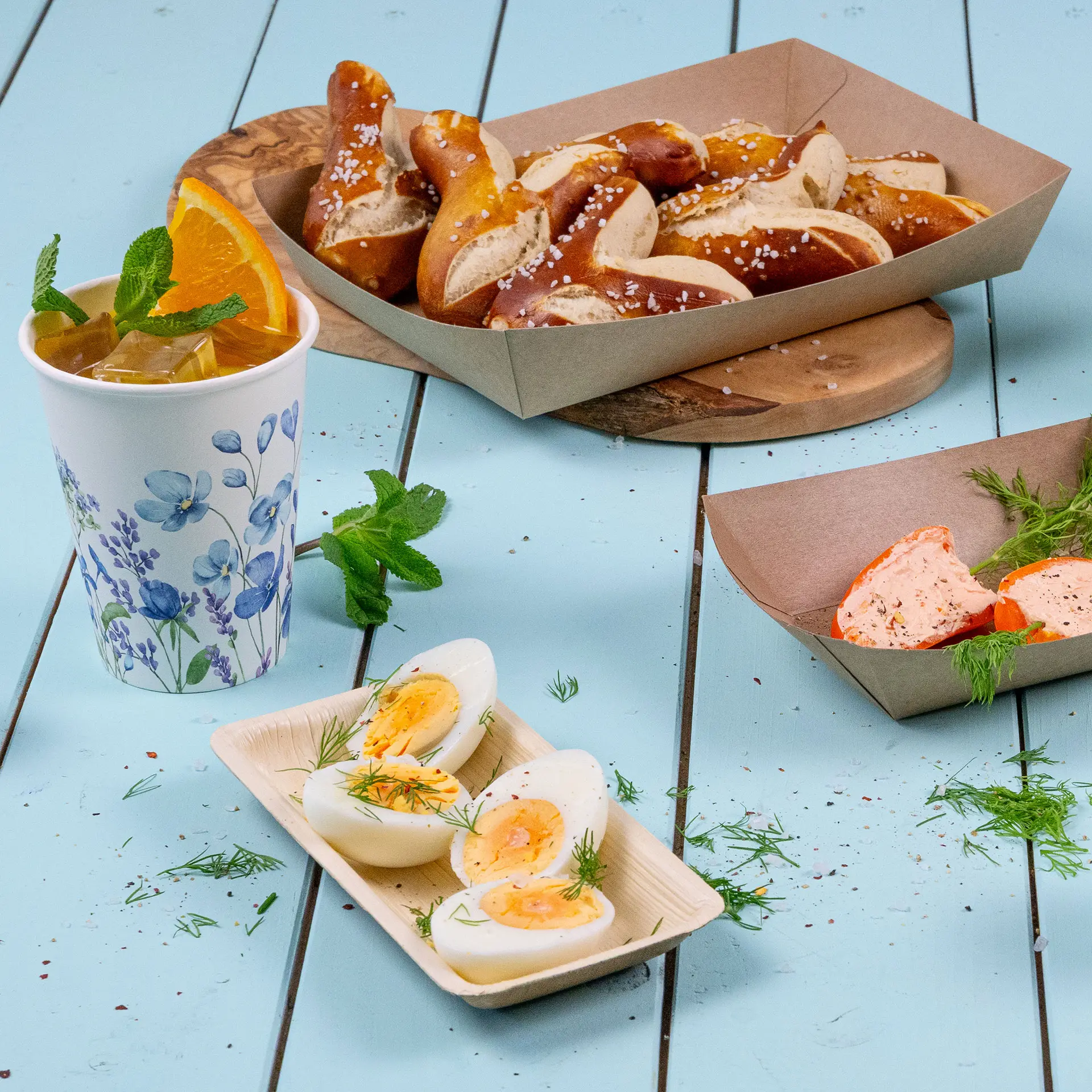 Una ciotola rettangolare in foglia di palma contenente uova sode, guarnite con aneto; una tazza di tè freddo con menta e arancia; pretzel in una scatola vicina.