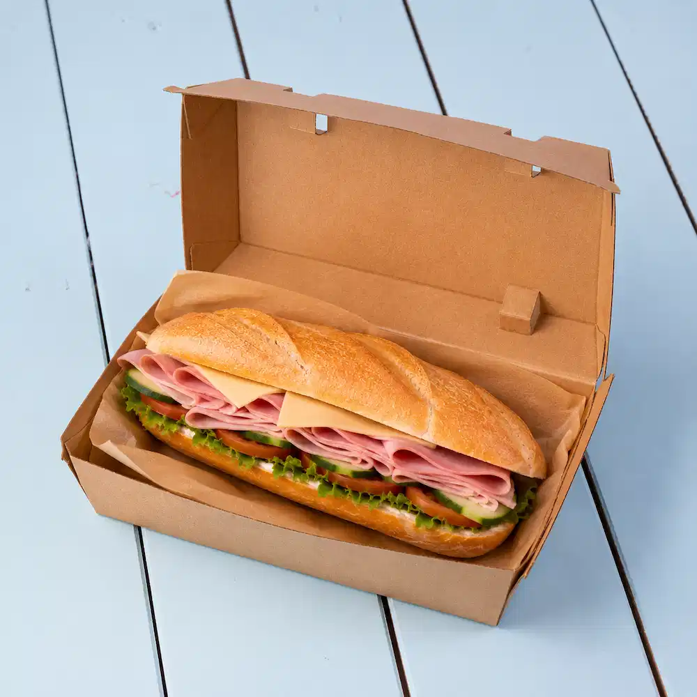 Panino in una scatola da asporto marrone, con strati di prosciutto, formaggio, lattuga e cetrioli affettati.