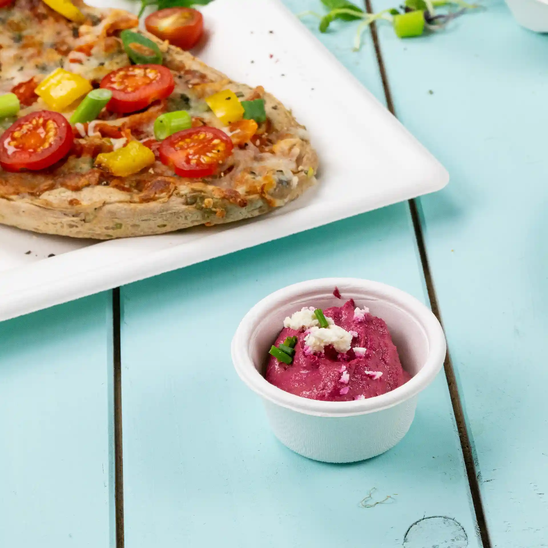 Ciotola di bagassa bianca con salsa rosa e guarnizione verde, posizionata accanto a una pizza su un piatto bianco.