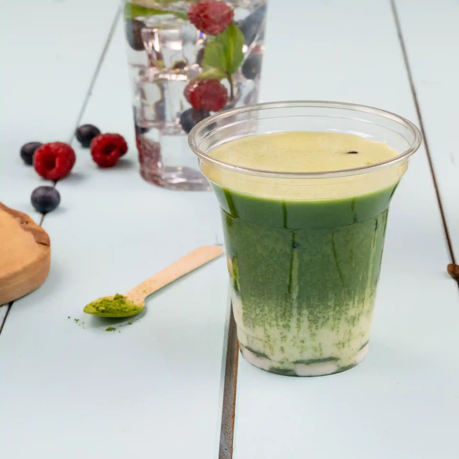Bicchiere rPET trasparente riempito di bevanda verde, con un cucchiaio di legno e bacche sullo sfondo.