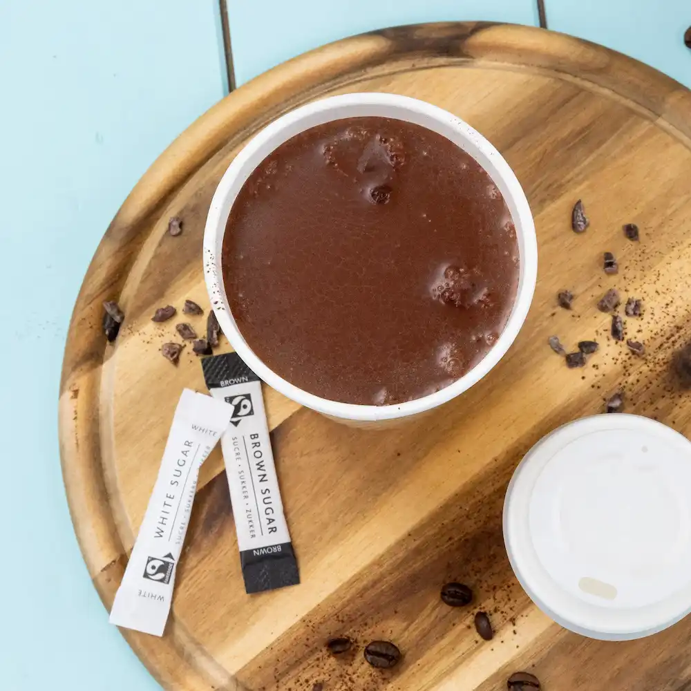 Cioccolata calda in una tazza bianca con bustine di zucchero, su un vassoio di legno con chicchi di caffè e cacao sparso.