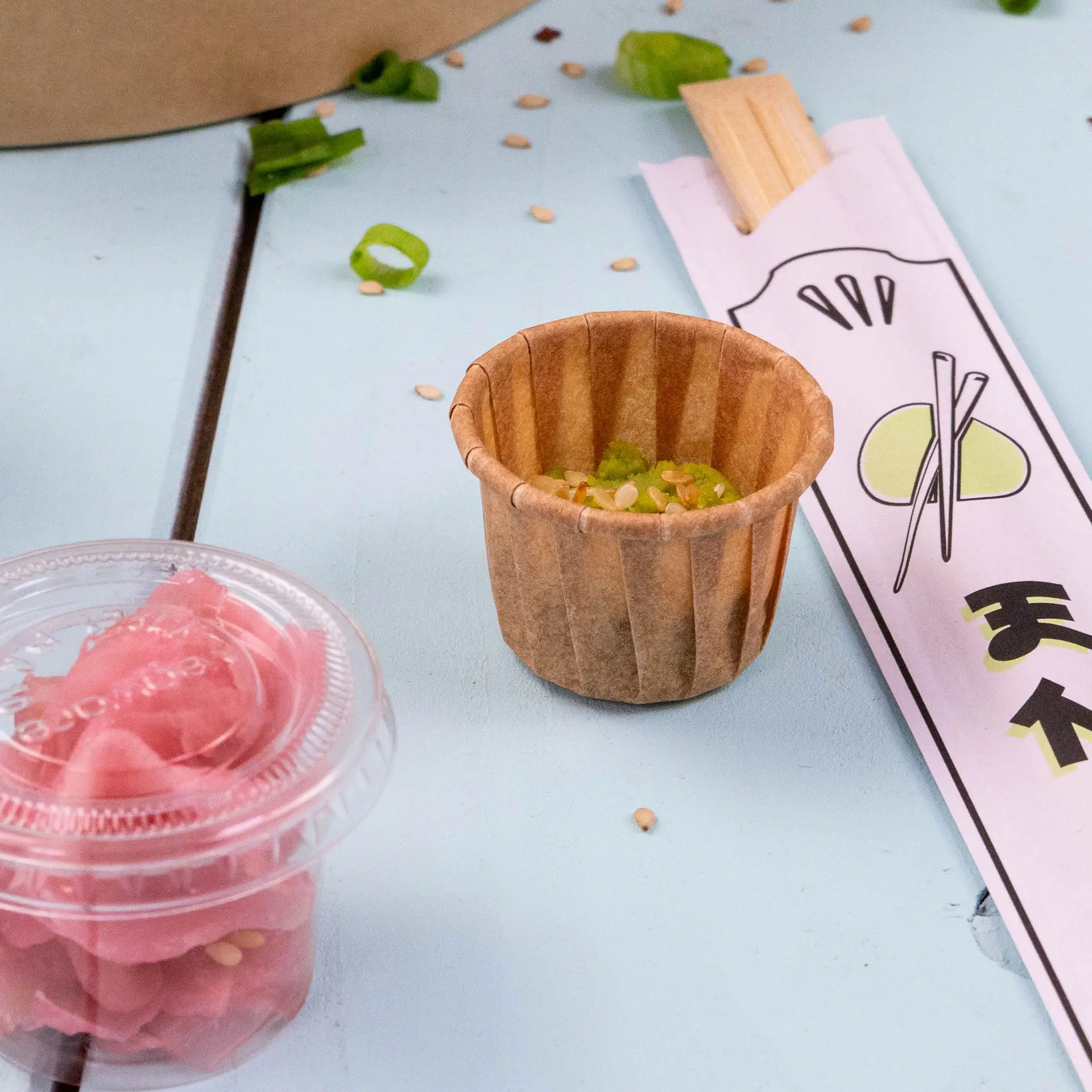 Tazza di carta marrone per salse e condimenti, capacità di 15 ml, con semi di sesamo e cipollotti tritati accanto a zenzero sottaceto e bacchette.