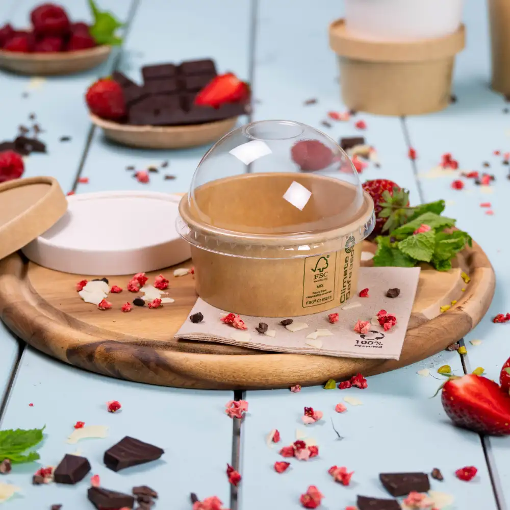 Contenitore per dessert da asporto con coperchio trasparente su un vassoio di legno, circondato da fragole fresche, lamponi e pezzi di cioccolato.