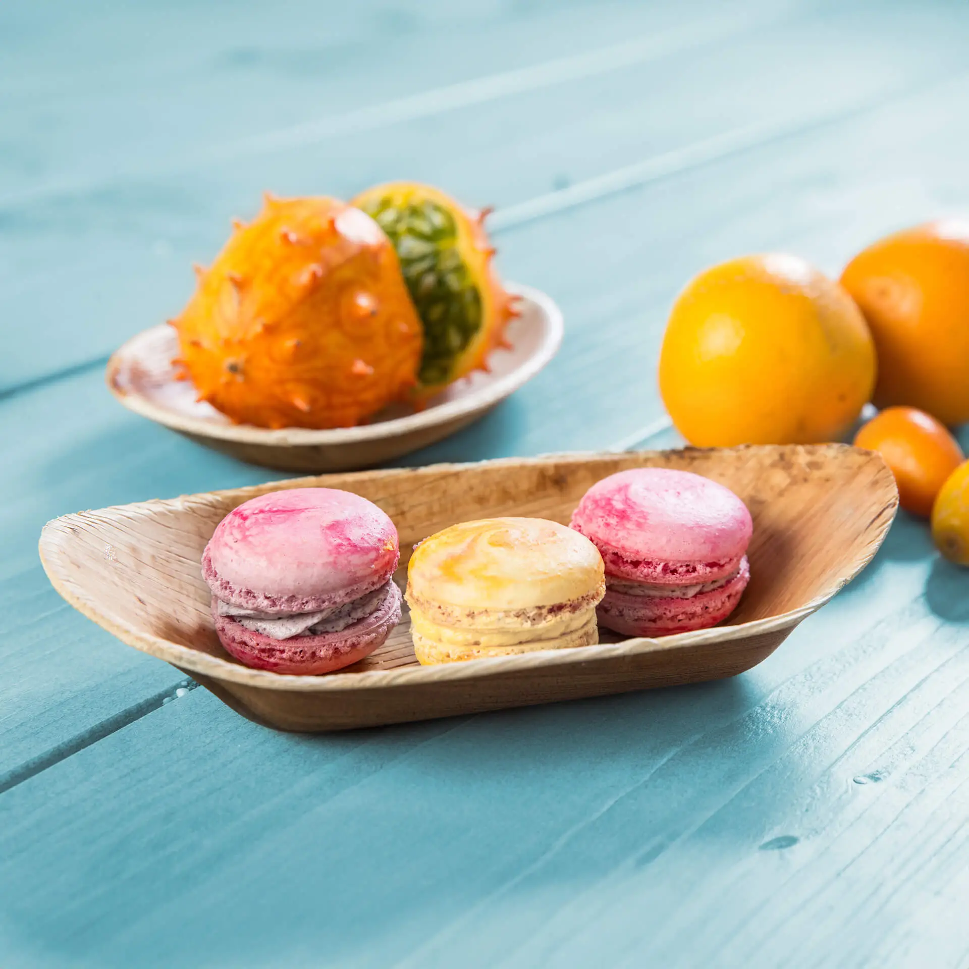 Tre macarons colorati disposti in una barca di foglie di palma, accanto a frutta fresca, su una superficie di legno blu.