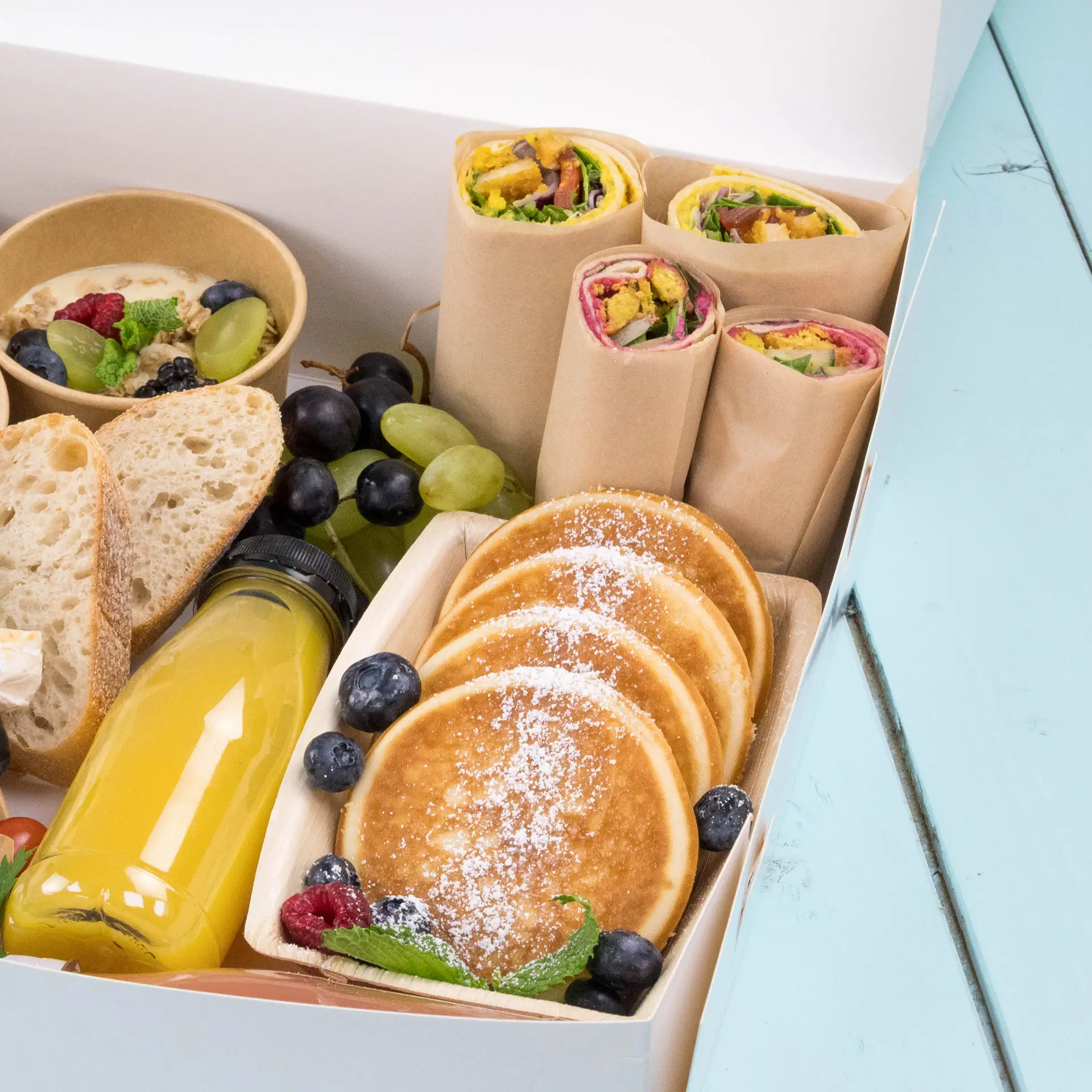 Una scatola piena di articoli per la colazione, tra cui wrap, pancake, frutta, pane e una bottiglia di succo.
