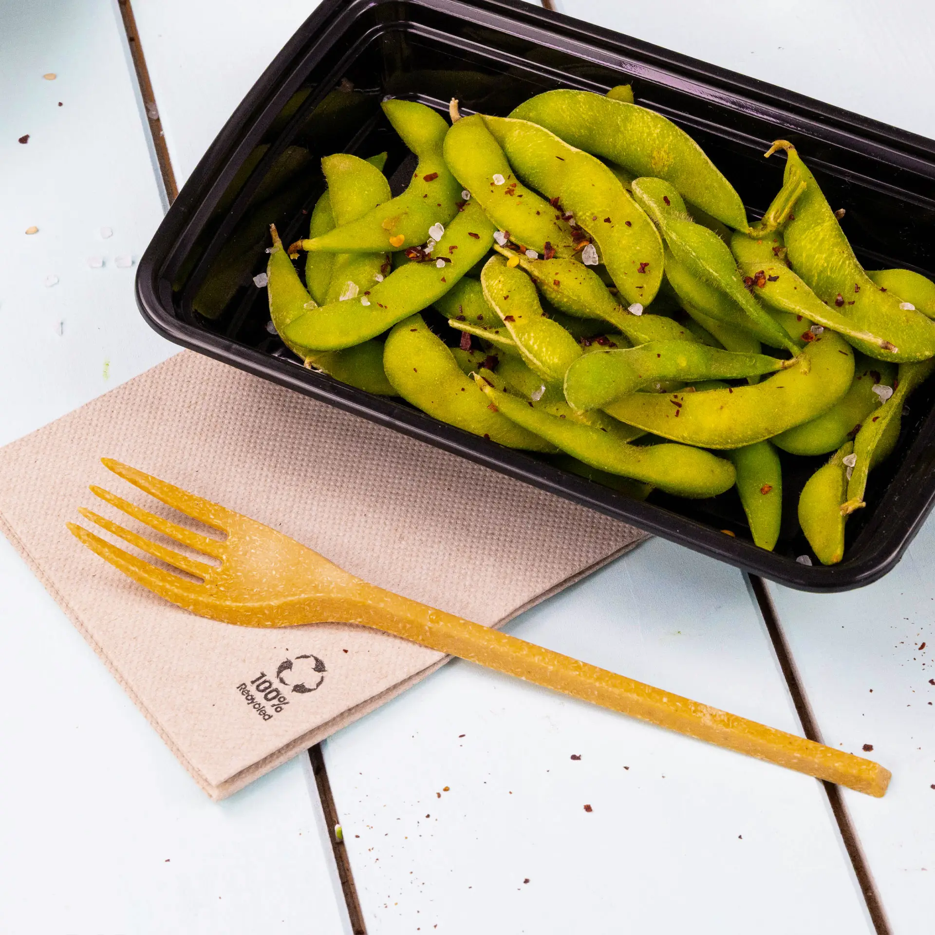 Immagine di fave edamame fresche in un contenitore nero, accompagnate da una forchetta biodegradabile su un tovagliolo di carta riciclata.