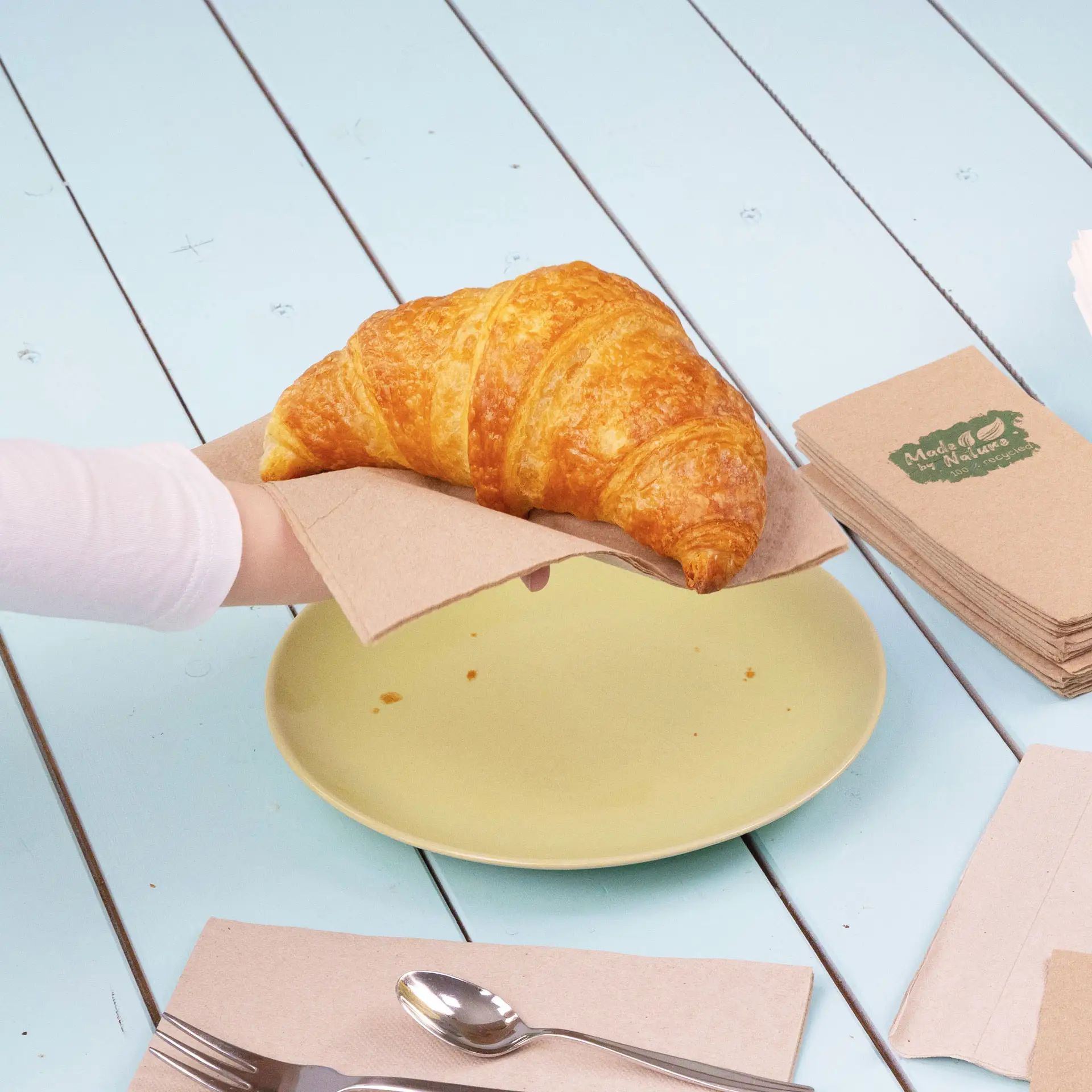 Una mano tiene un croissant su un tovagliolo di carta sopra un piatto giallo, con tovaglioli e posate vicini.