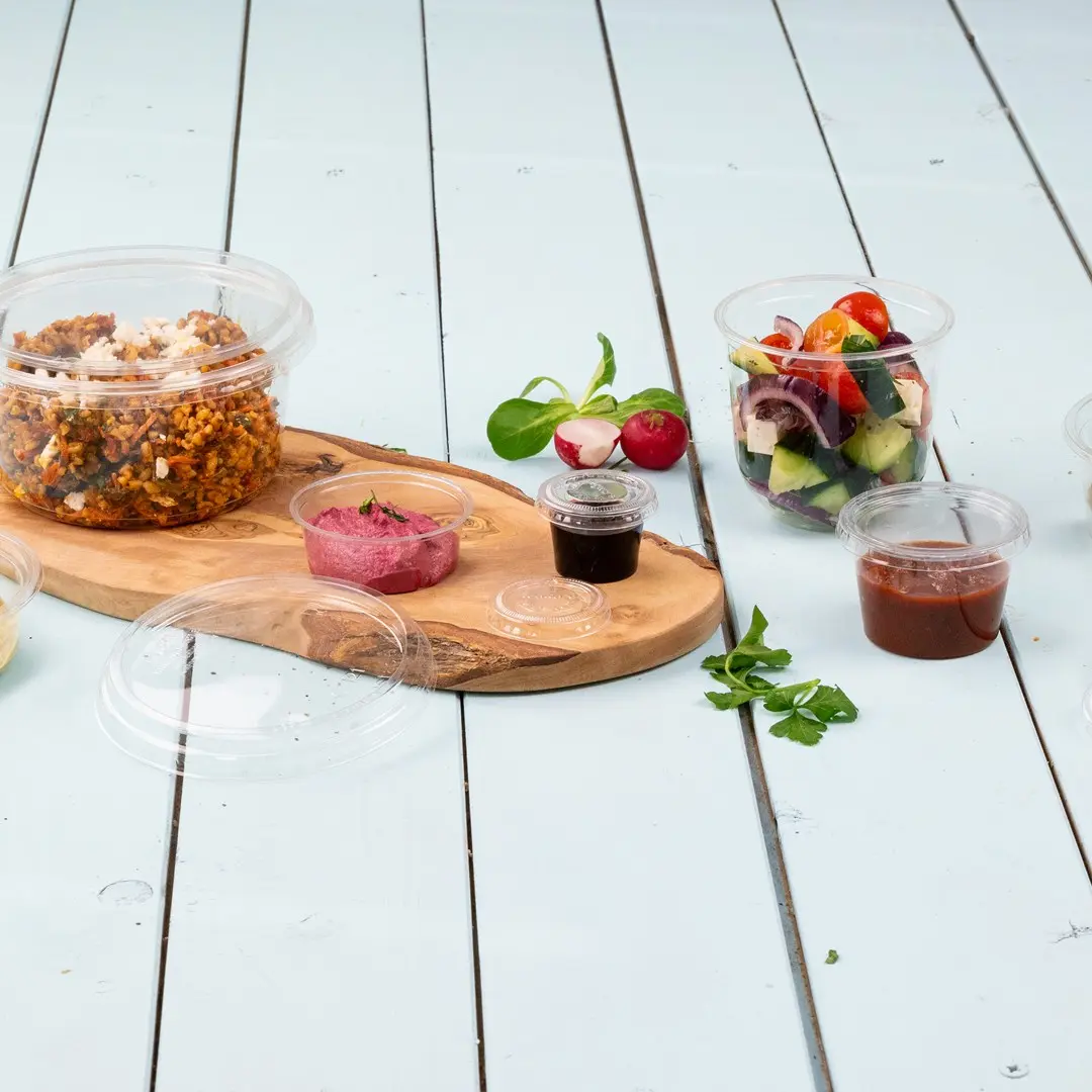 Contenitori in plastica trasparente con vari alimenti, tra cui un'insalata, salse e un piatto di cereali, disposti su un tagliere di legno.