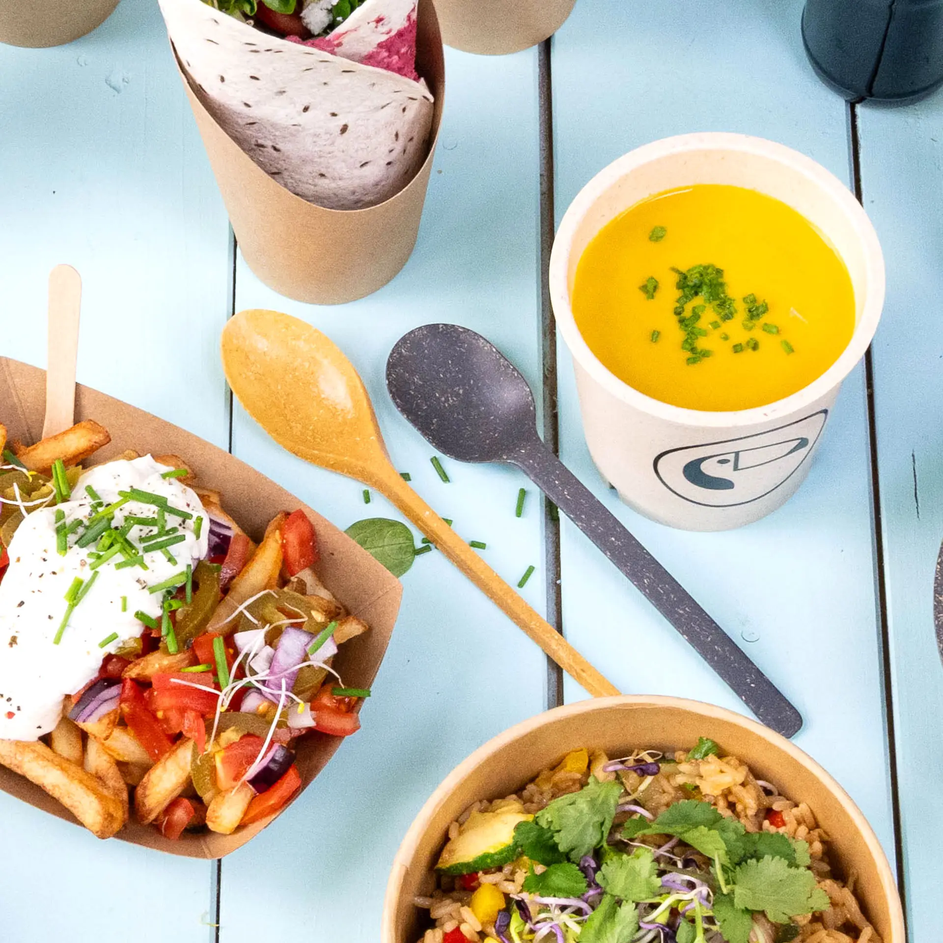 Un cucchiaio riutilizzabile marrone caramello realizzato in biocomposito; include vari alimenti e una tazza gialla per zuppa con erba cipollina.