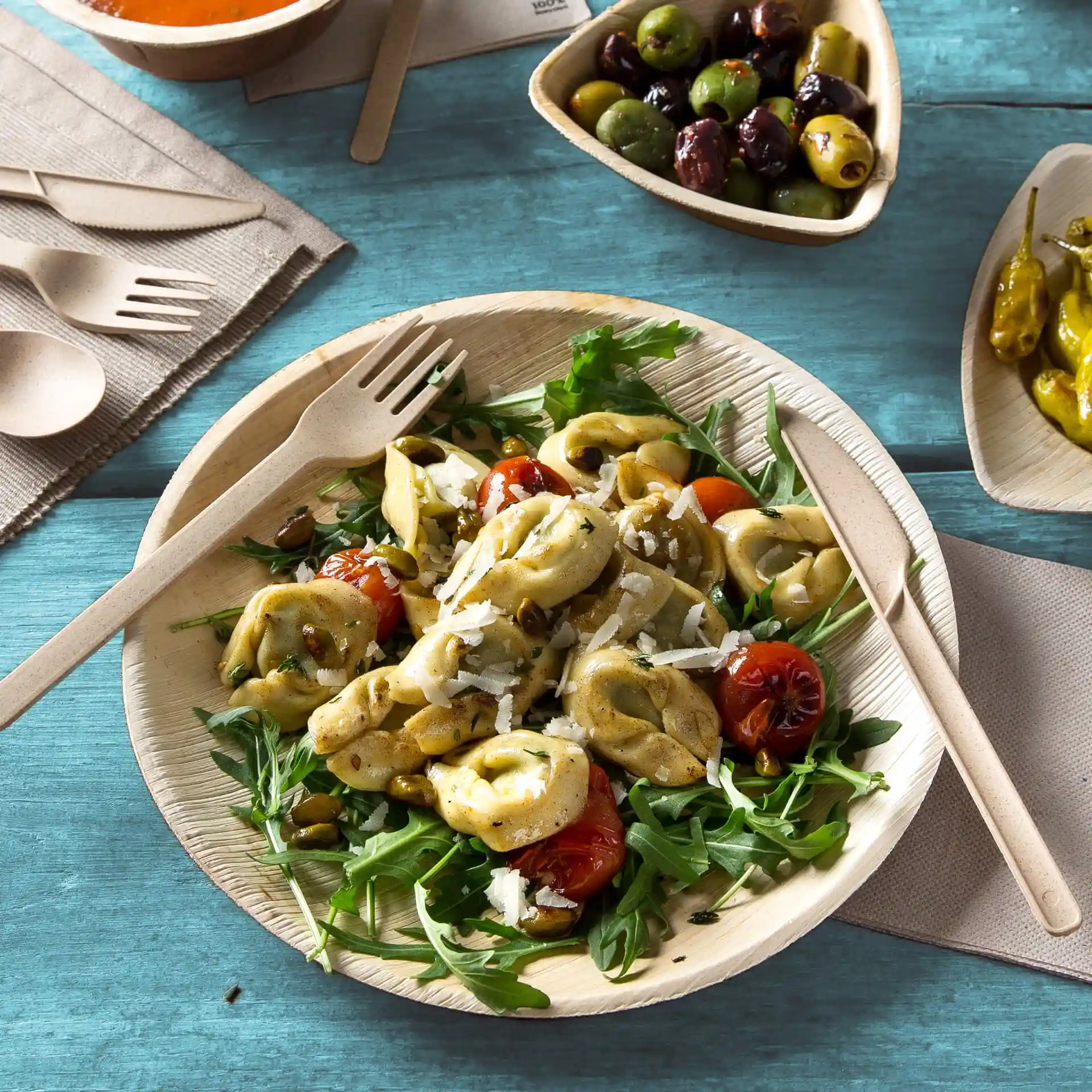 Piatto rotondo in foglia di palma con insalata, tortellini e pomodorini, accompagnato da posate di legno su una superficie blu.