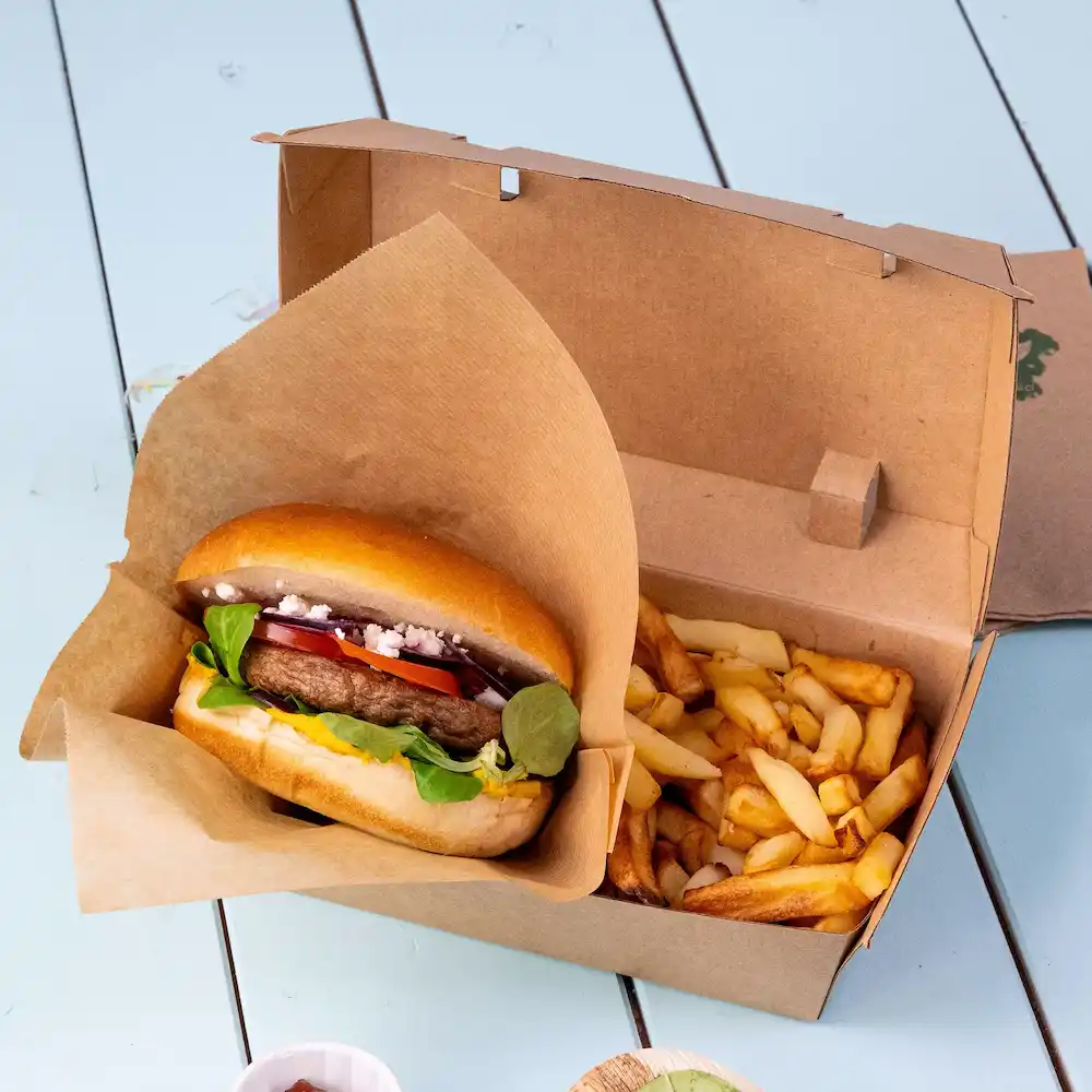 Burger in involucro di carta con verdure fresche accanto a una scatola di patatine su una superficie blu.