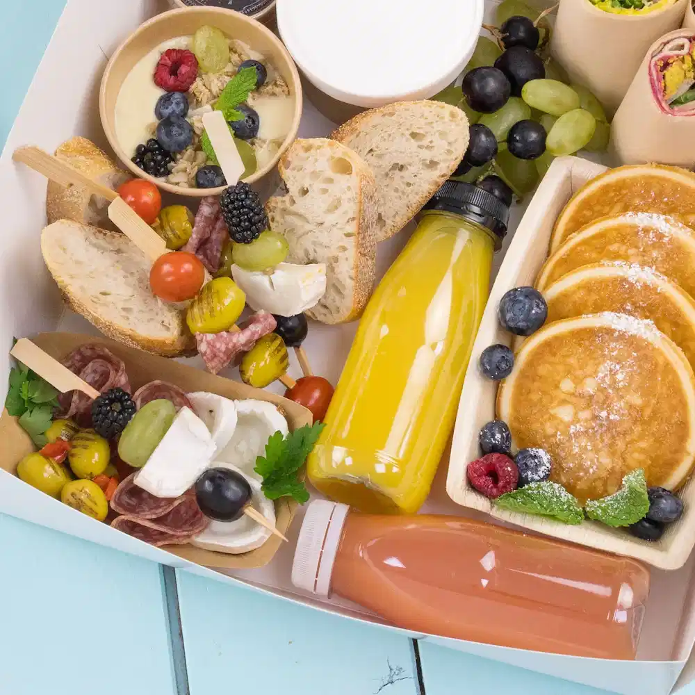 Una varietà di cibi tra cui pancake, frutta, pane e bevande, esposti in un contenitore su una superficie azzurro chiaro.