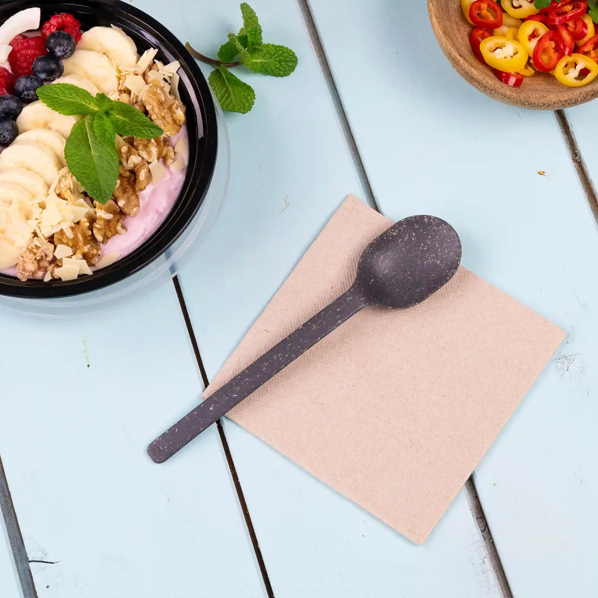 Immagine di un cucchiaio nero su un tovagliolo quadrato non sbiancato, con una ciotola di yogurt guarnita con frutta e noci sullo sfondo.