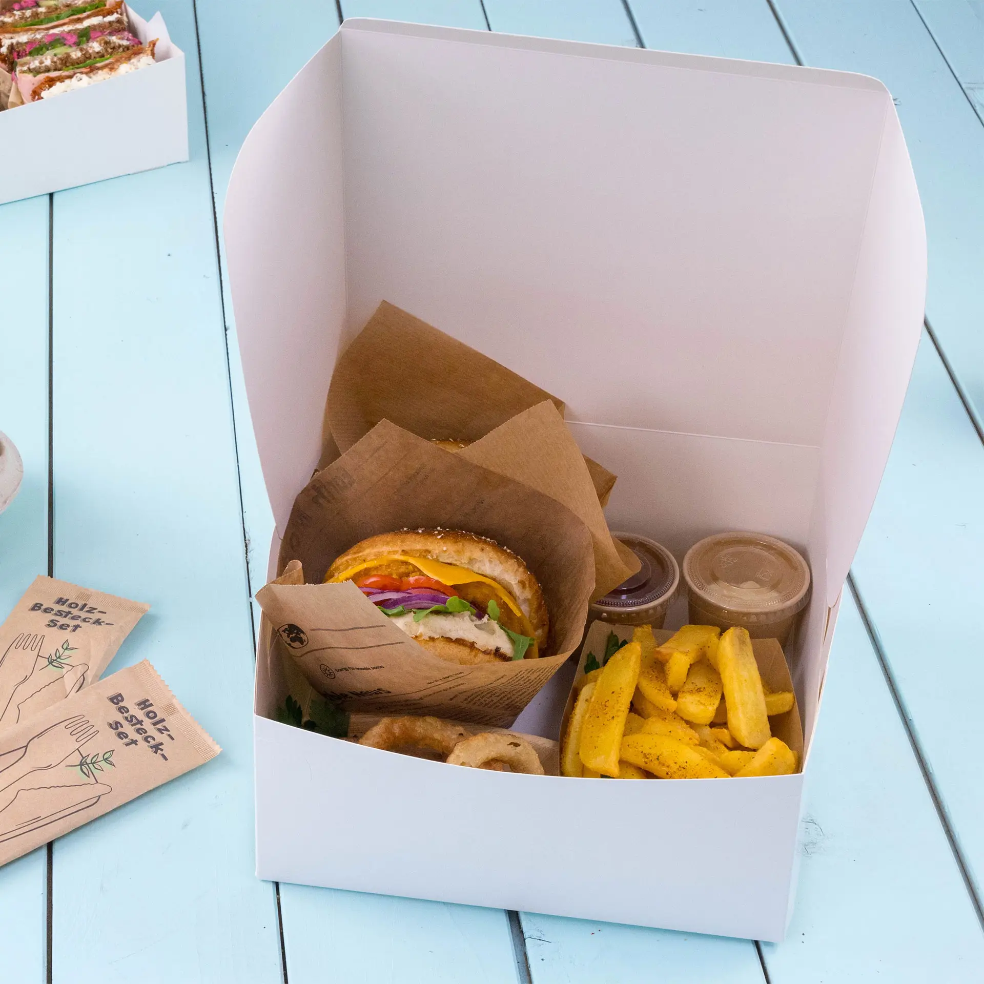 Scatola da asporto contenente un panino, patatine e due piccoli contenitori, posizionata su una superficie azzurro chiaro.