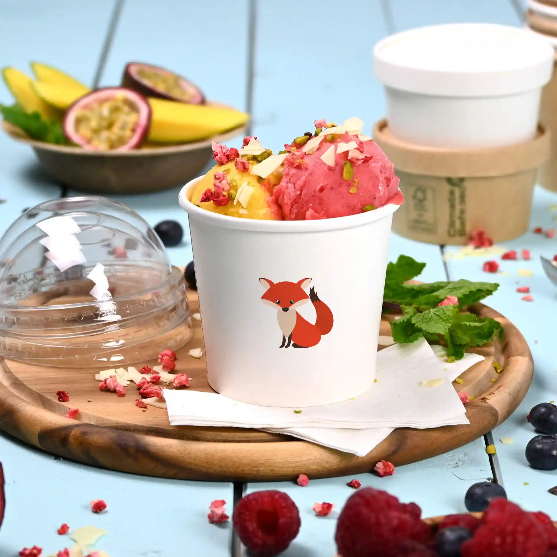 Coppetta di cartone bianca per gelato con illustrazione di volpe, piena di gelato rosa e giallo, su un piatto di legno.