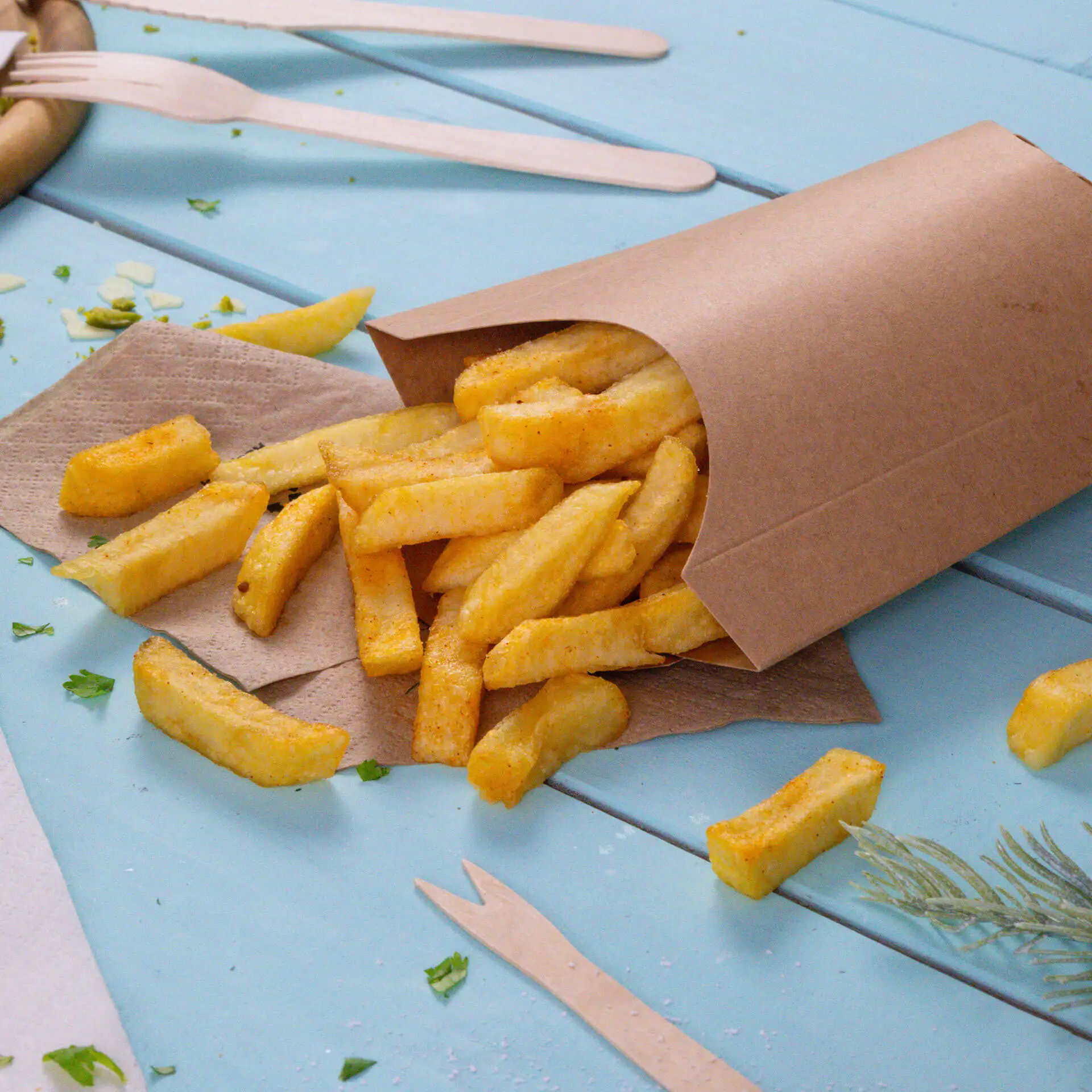 Cucchiaio in cartone marrone con patatine che traboccano, posizionato su una superficie blu con posate di legno nelle vicinanze.