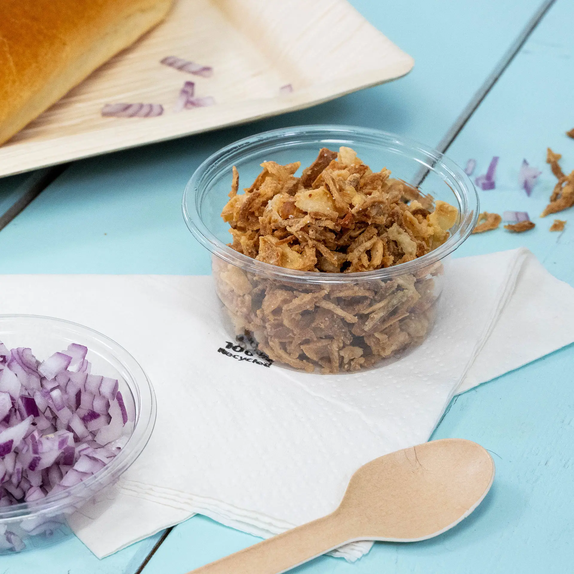 Un bicchiere trasparente con cipolle fritte croccanti è posizionato accanto a cipolle rosse tritate su un tovagliolo, con un cucchiaio di legno vicino.