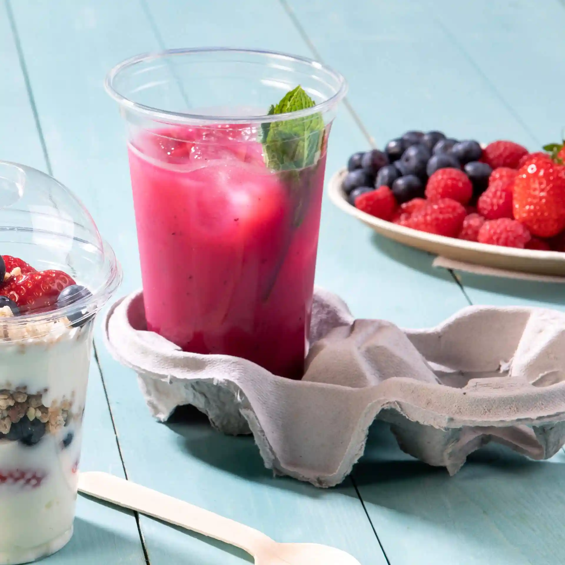 Bicchiere rPET trasparente da 16 oz riempito con bevanda rosa e menta, posizionato in un portauova grigio con un parfait di yogurt vicino.