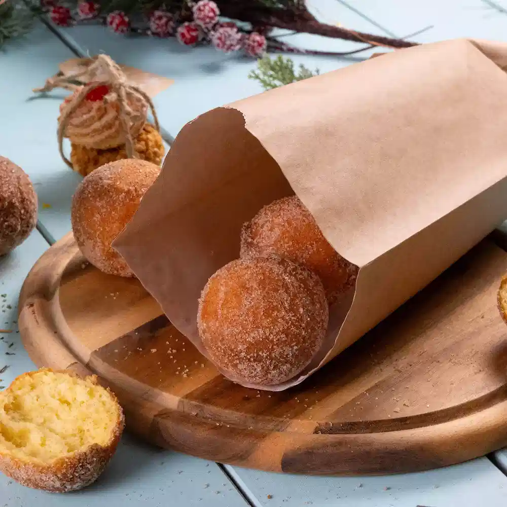 Una busta di carta marrone contiene diversi ciambelle rotonde cosparse di zucchero a velo su un tagliere di legno, con una ciambella parzialmente tagliata.