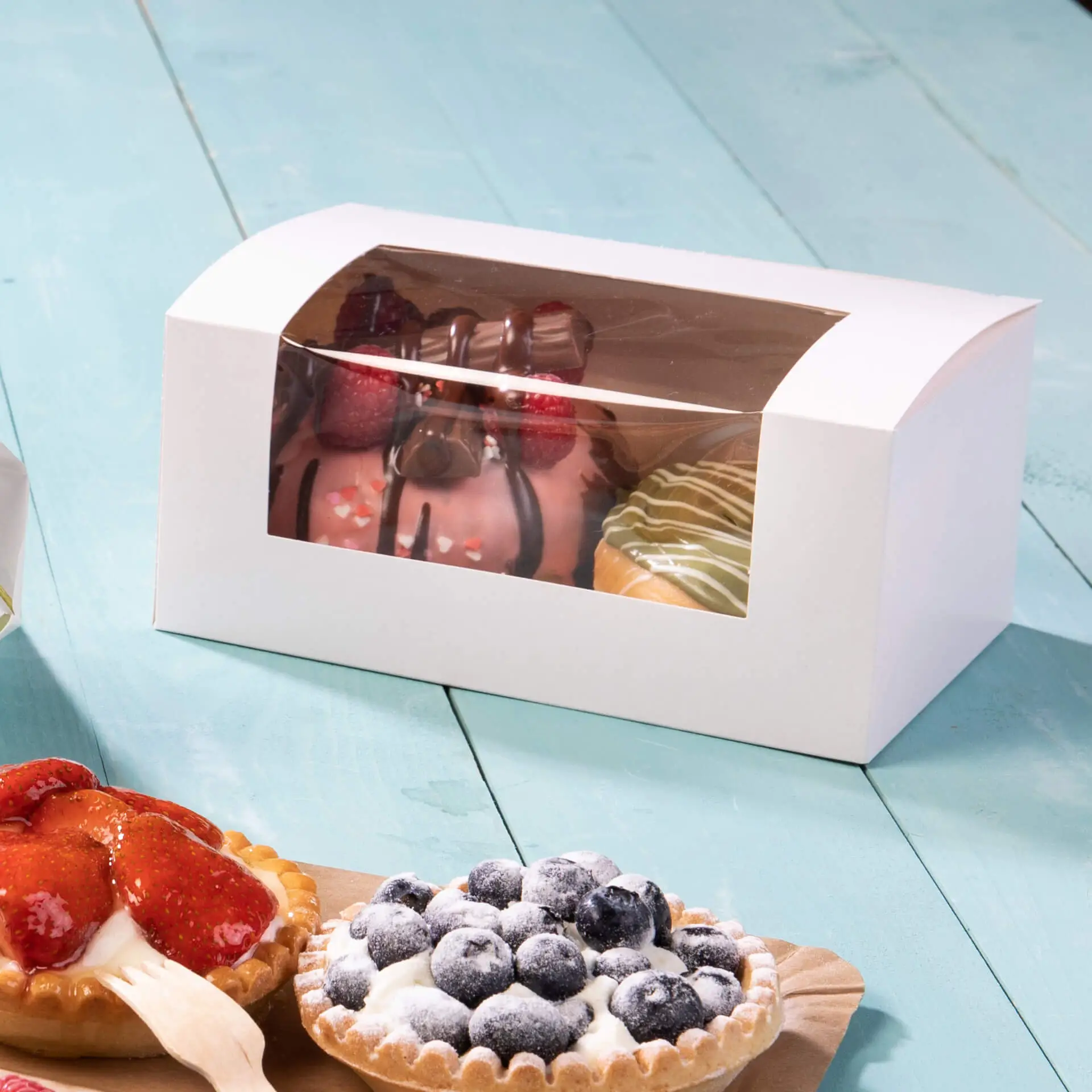 Scatola bianca per cupcake con una finestra in cellulosa che mostra pasticcini assortiti, tra cui torte e crostate, su una superficie turchese.