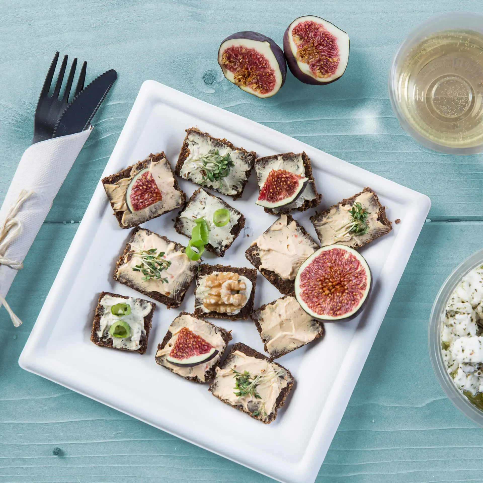 Un piatto quadrato bianco in canna da zucchero tiene diversi canapés guarniti con fichi, noci ed erbe; posate nere accanto.