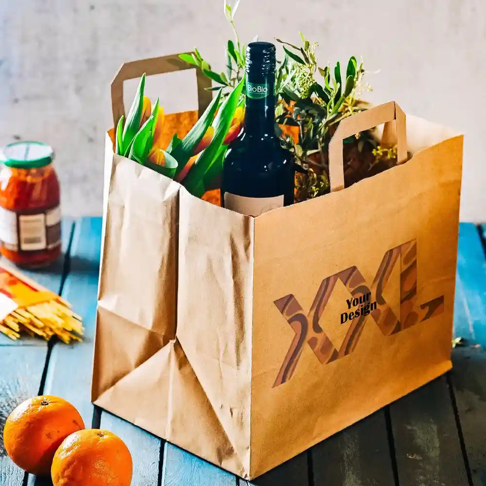 Busta di carta marrone con manici, contenente una bottiglia di vino, fiori e un barattolo, su una superficie di legno con arance.