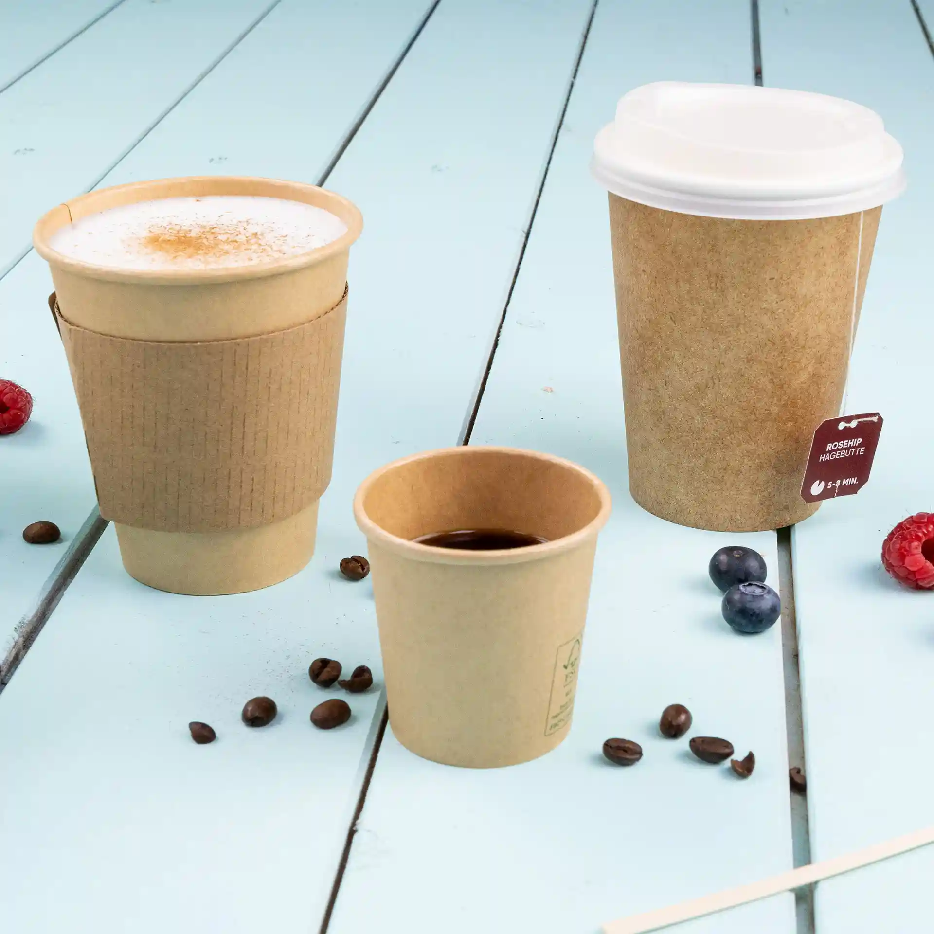 Immagine di tre bicchieri di carta marroni di diverse dimensioni su una superficie di legno blu, con chicchi di caffè e bacche sparsi.