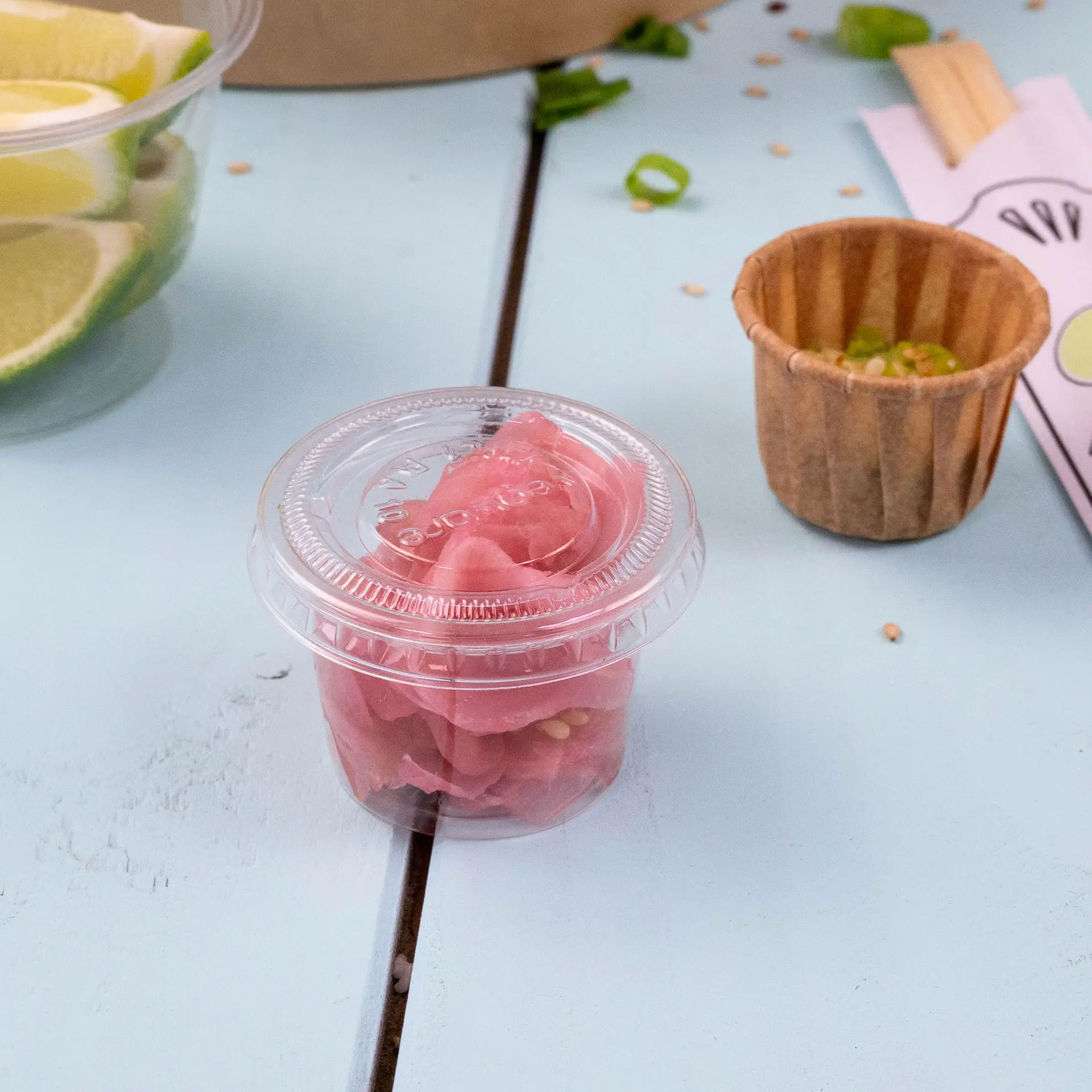 Un bicchiere di plastica trasparente contenente zenzero marinato rosa su una superficie azzurra, con un bicchiere di carta marrone e fette di lime sullo sfondo.