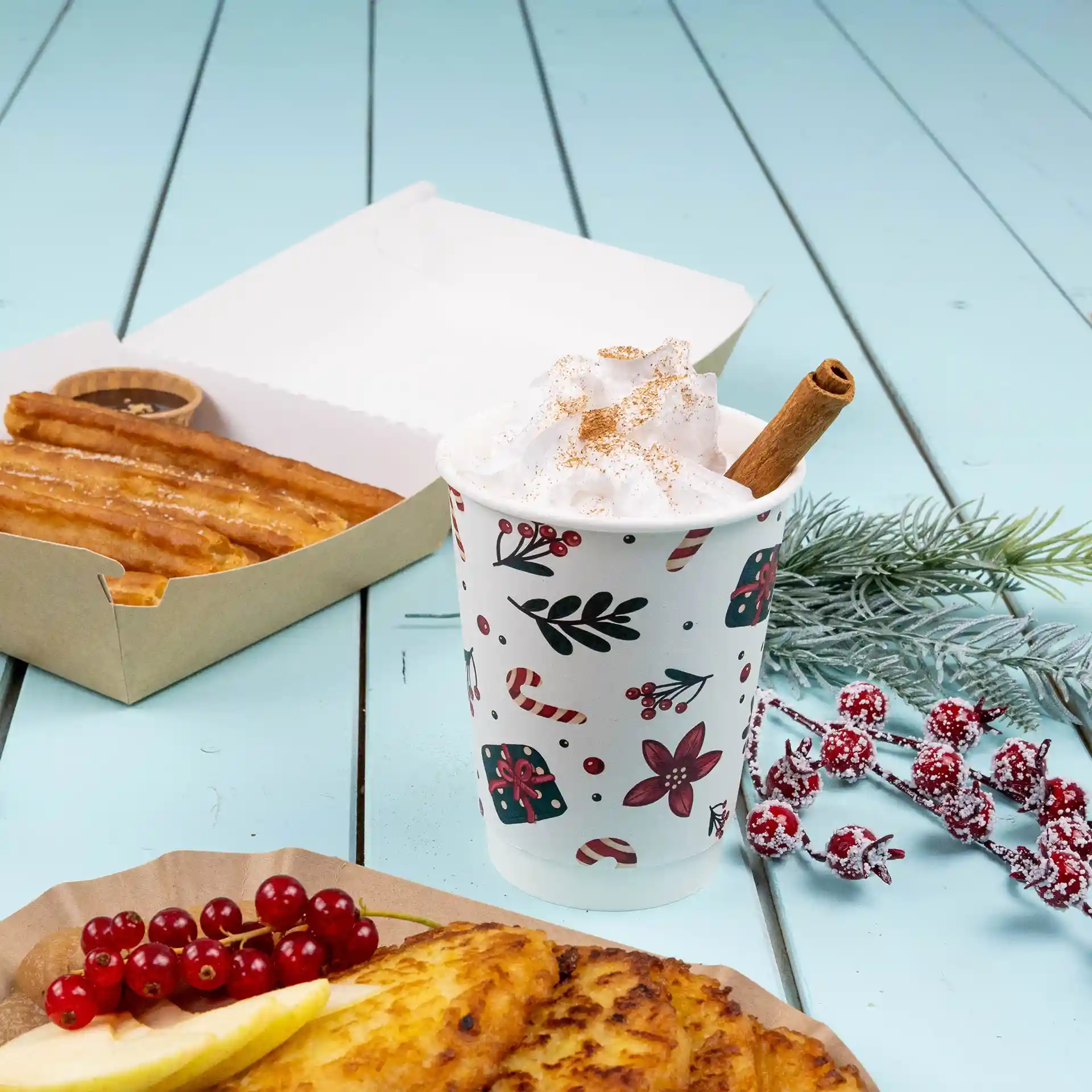 Tazza a doppia parete con design invernale, riempita di panna montata e un bastoncino di cannella, accanto a churros e frutta.