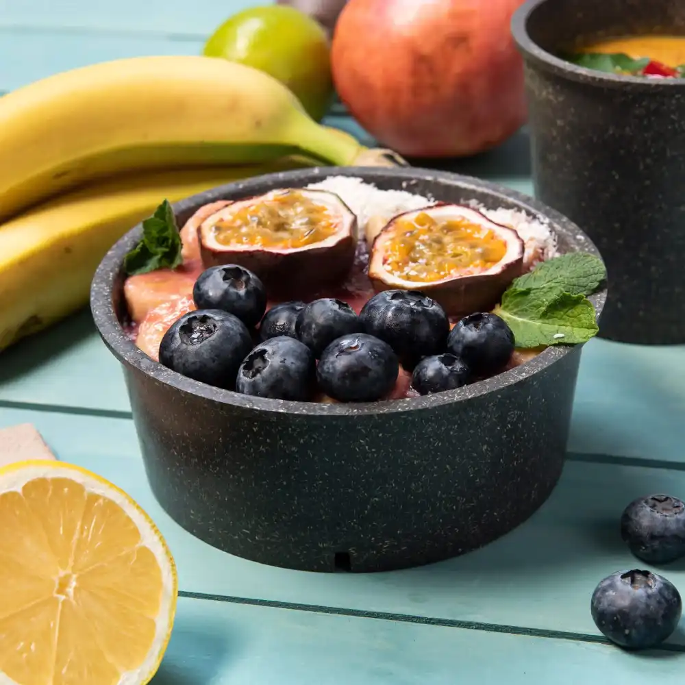 Una ciotola nera piena di frutta, comprese le mirtilli e frutto della passione, circondata da banane e una fetta di limone.