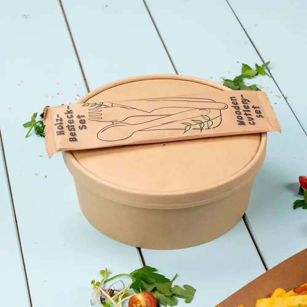 Contenitore marrone con coperchio e un set di posate in legno confezionato sopra, con erbe verdi e elementi di insalata vicini.