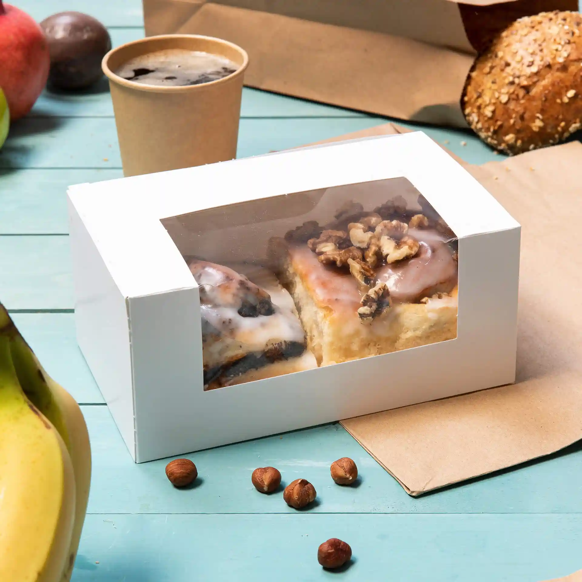 Scatola di pasticceria bianca con una finestra in cellulosa contenente due dolci, circondata da una banana, nocciole e una tazza.
