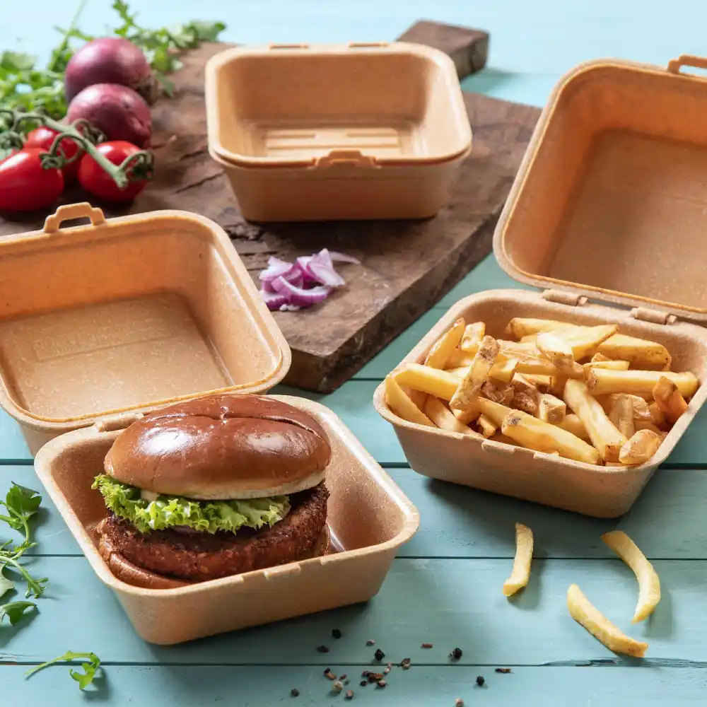 Contenitore con un hamburger con lattuga, accanto a un contenitore con patatine, su una superficie di legno con verdure vicine.