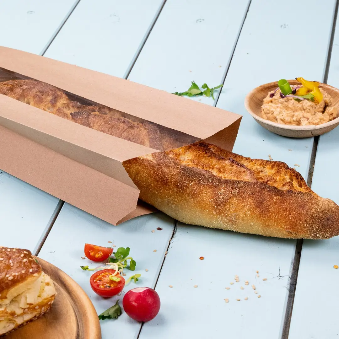 Busta di carta marrone parzialmente aperta con una baguette, pomodorini, ravanelli e una ciotola di salsa su una superficie azzurro chiaro.