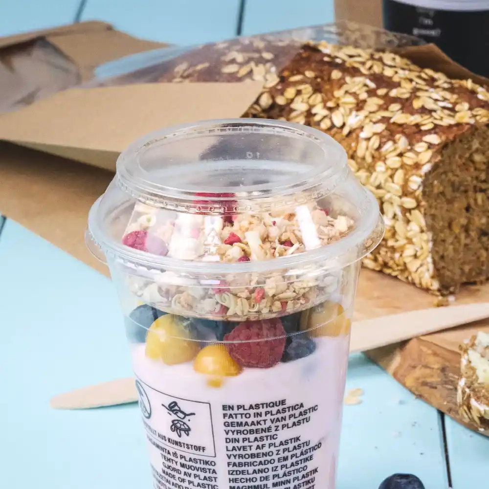 Tazza con yogurt, granola e bacche, accanto a un pane d'avena su una superficie blu.