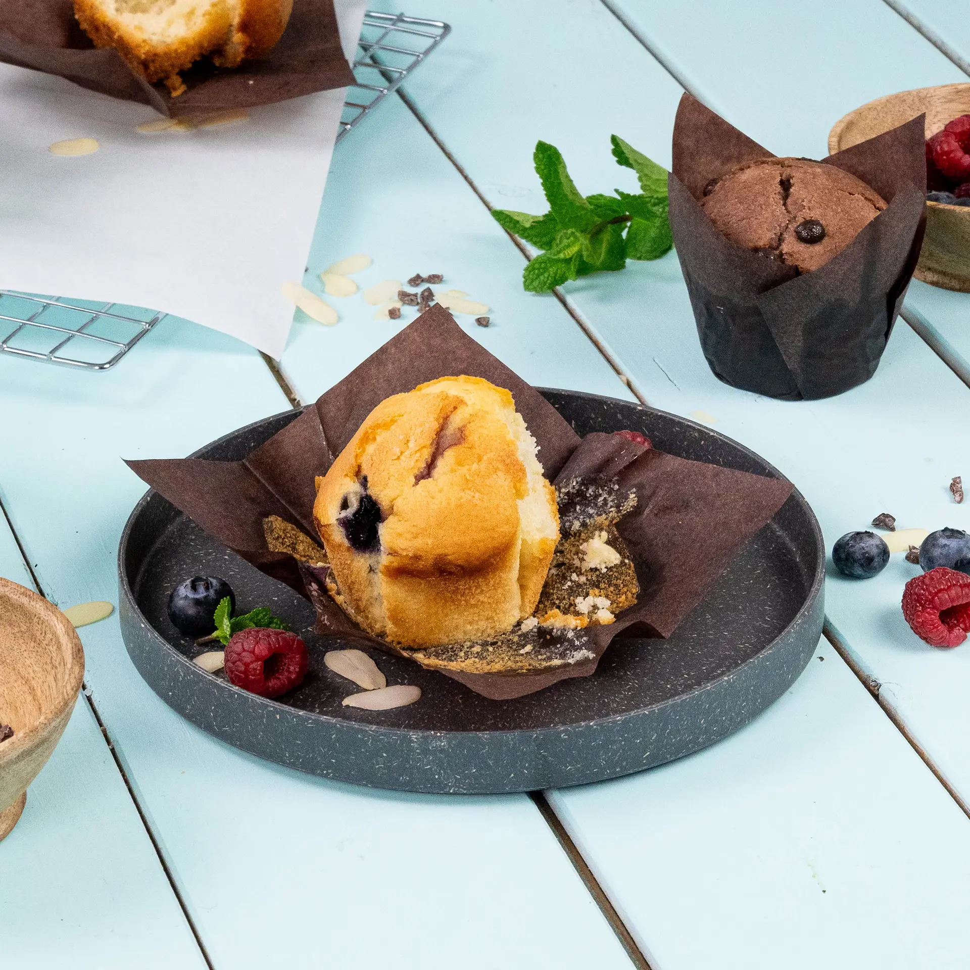 Muffin in un cup di carta marrone, con mirtilli e lamponi su un piatto grigio, su uno sfondo azzurro chiaro.