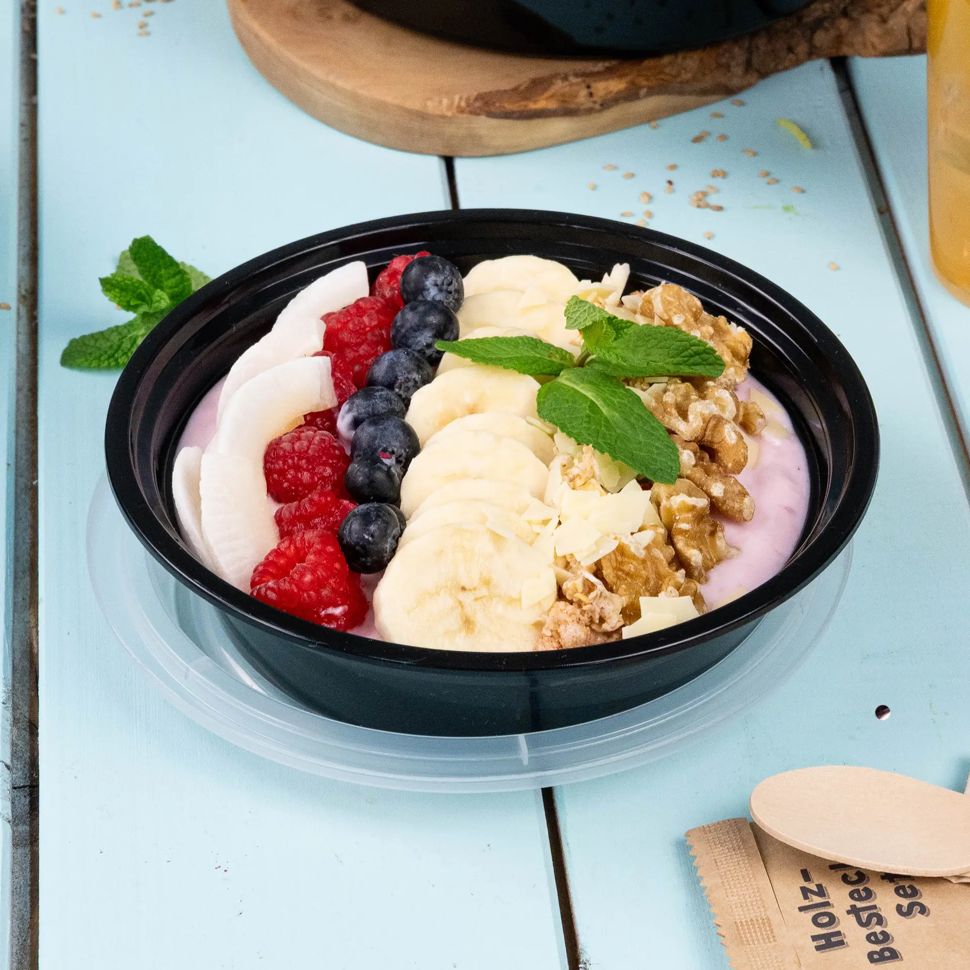 Un piatto nero riutilizzabile pieno di yogurt, fette di banana, mirtilli, lamponi e granola, guarnito con menta.