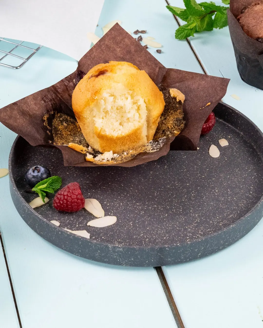Muffin in un involucro di carta marrone su un piatto scuro, circondato da lamponi, mirtilli, foglie di menta e fette di mandorla.