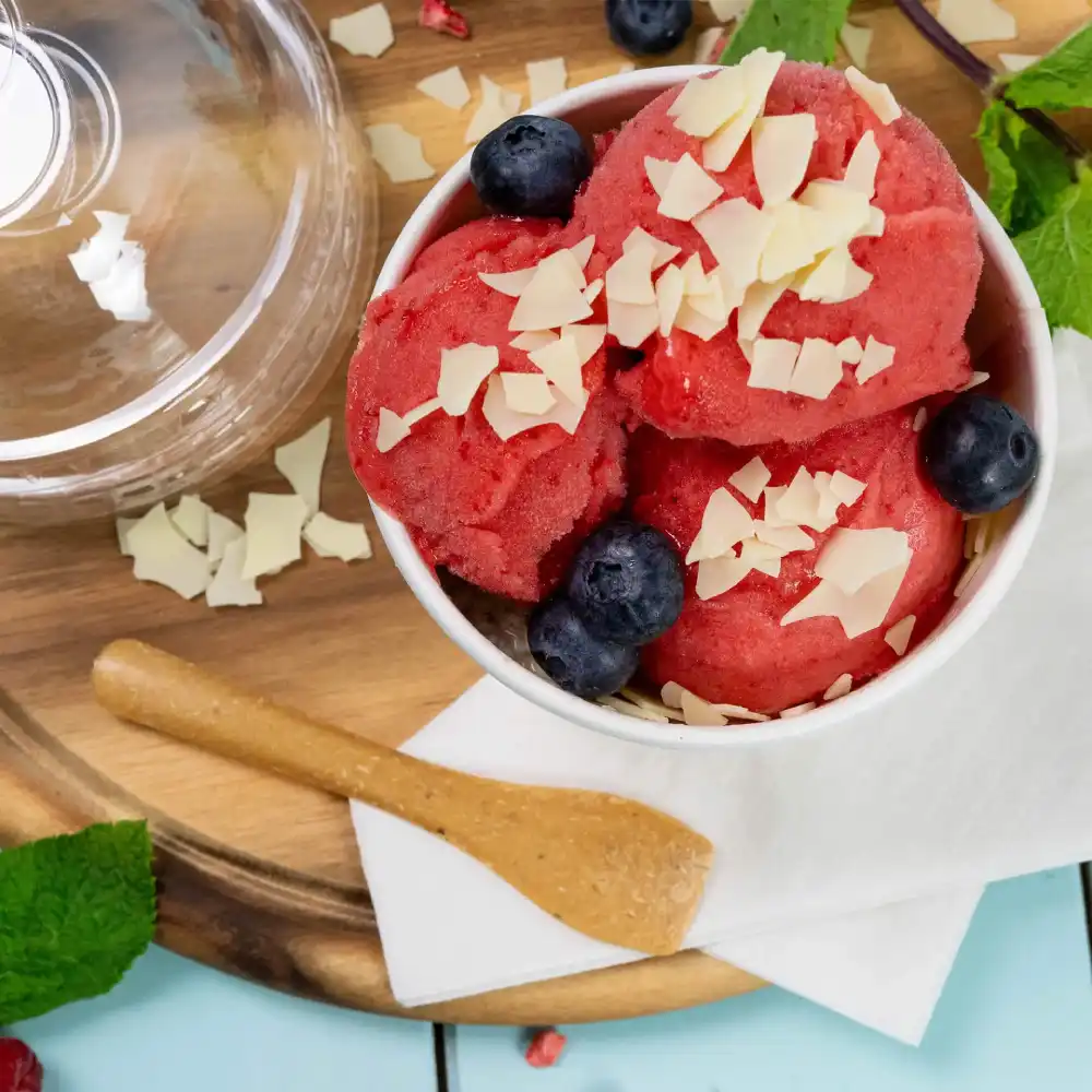 Ciotola di sorbetto rosso guarnito con scaglie di cioccolato e mirtilli, accompagnato da un cucchiaio di legno su un vassoio di legno.