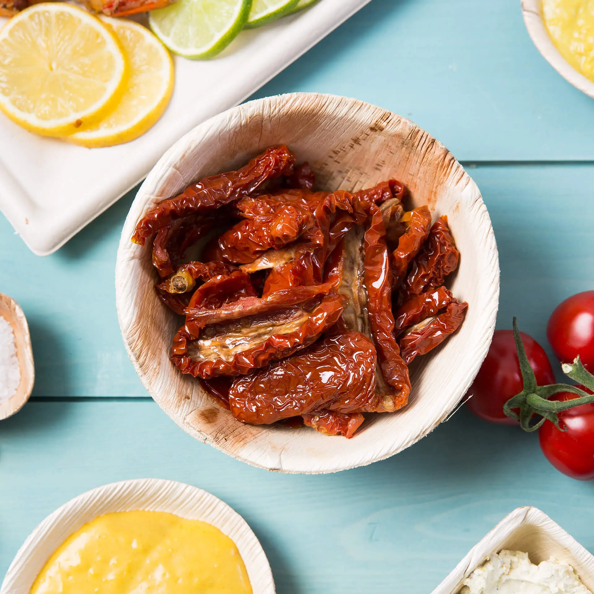 Pomodori secchi in una ciotola di foglie di palma su una superficie azzurra, con limone, lime e pomodorini nelle vicinanze.