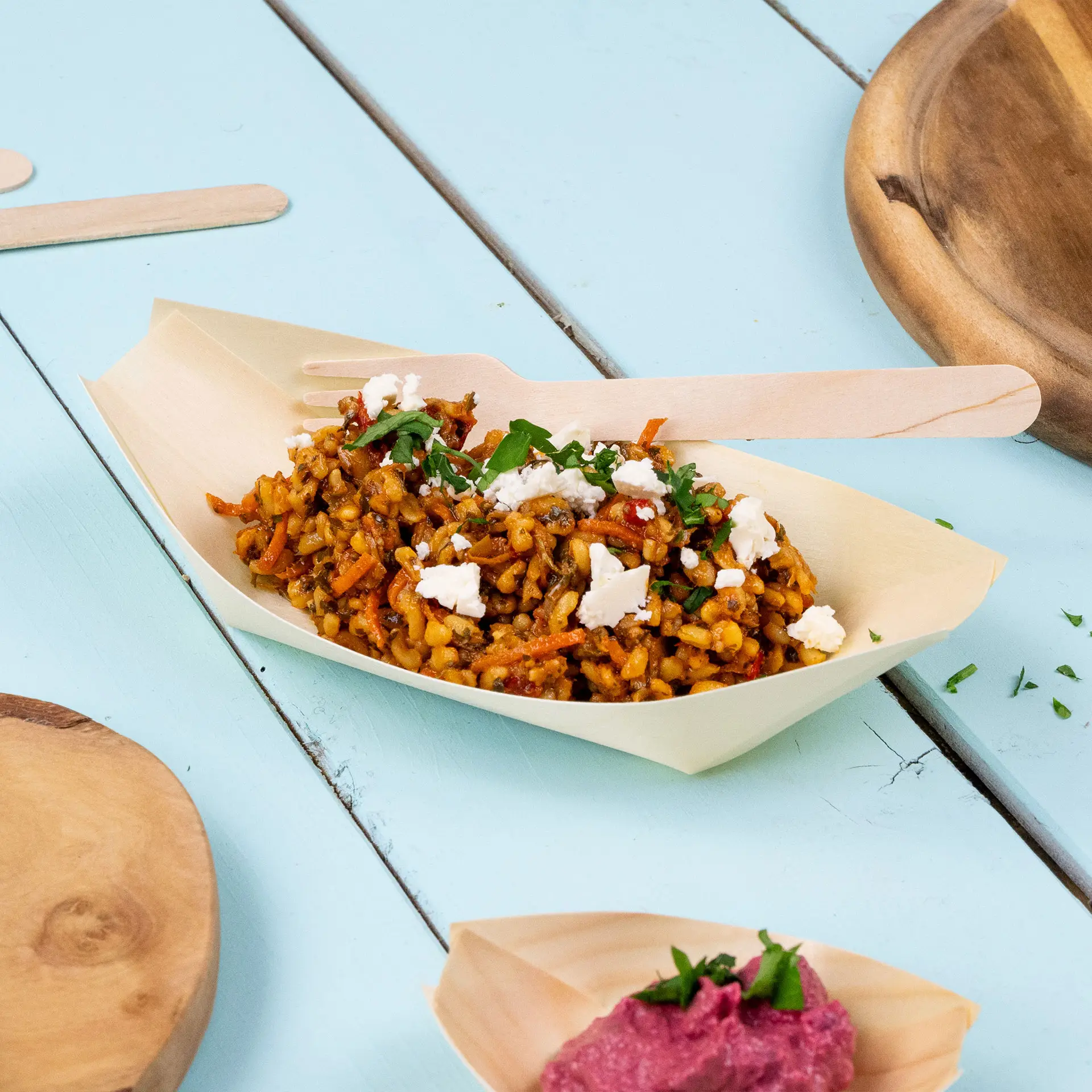 Una barca di legno piena di riso condito, guarnita con erbe e formaggio, posata su una superficie azzurro chiaro.