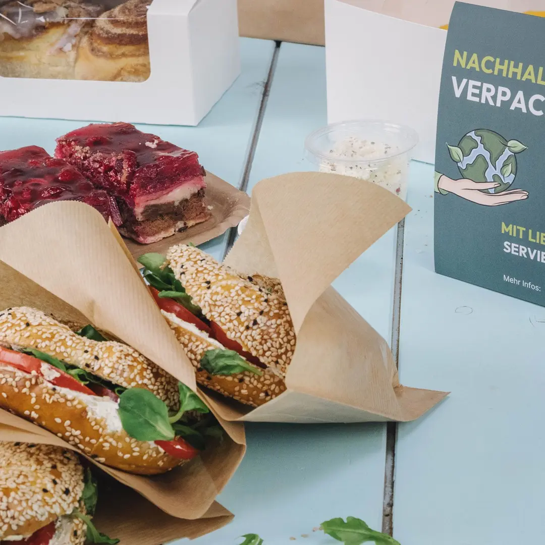 Tre panini ai semi di sesamo farciti con lattuga e pomodoro, un dessert con gelatina e un cartello sulla sostenibilità su un tavolo blu.