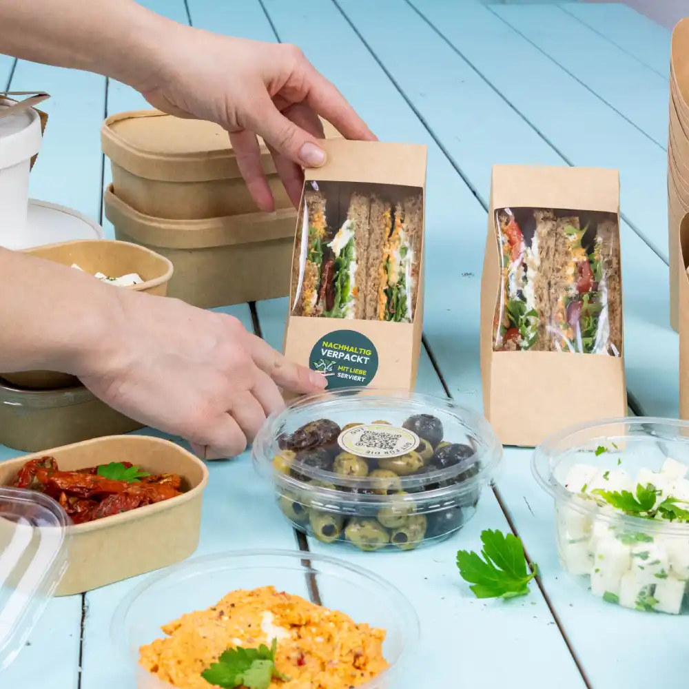 Una mano si avvicina a un pacchetto di sandwich su un tavolo; sono visibili vari contenitori per cibo, tra cui olive, un piatto cremoso e pomodori secchi al sole.