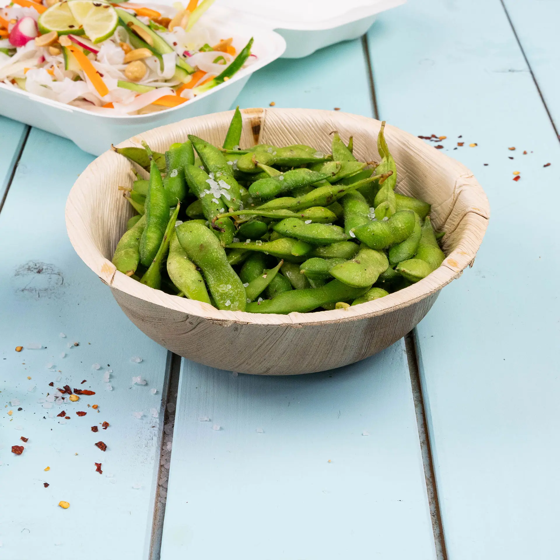 Ciotola riempita di fagioli edamame verdi con sale marino, posata su una superficie azzurra accanto a un'insalata.