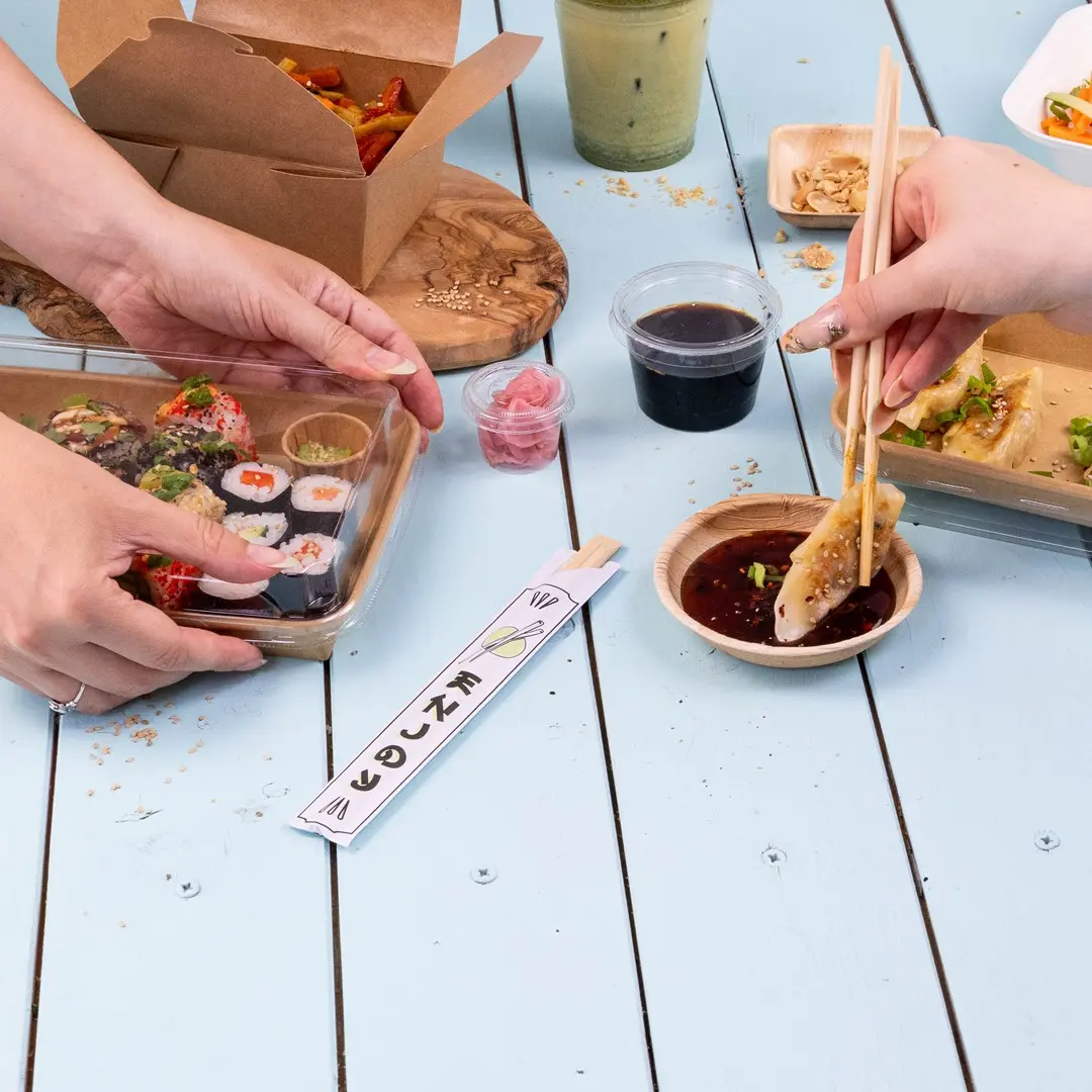 Mani che tengono sushi in un contenitore trasparente, con salsa, zenzero marinato e bacchette su un tavolo blu.