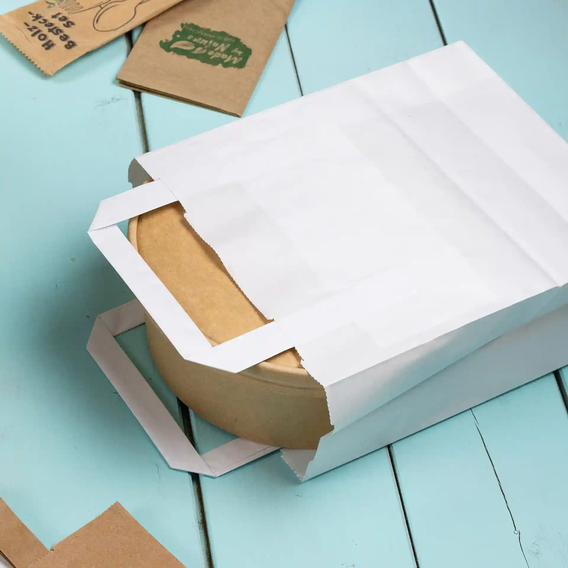 Borsa in carta kraft bianca parzialmente aperta, con un contenitore rotondo marrone all'interno, su una superficie azzurro chiaro.