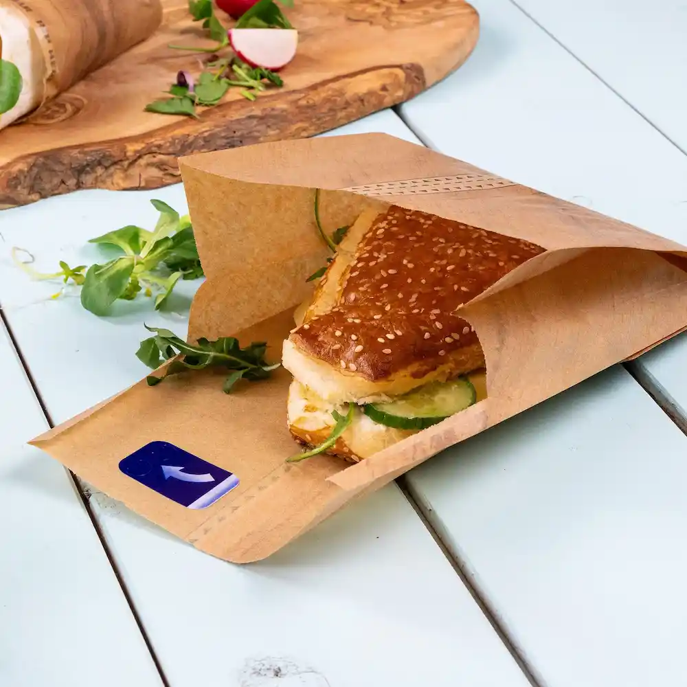 Sacchetto di carta marrone con un panino ai semi di sesamo e cetrioli, circondato da verdure fresche su una superficie blu.