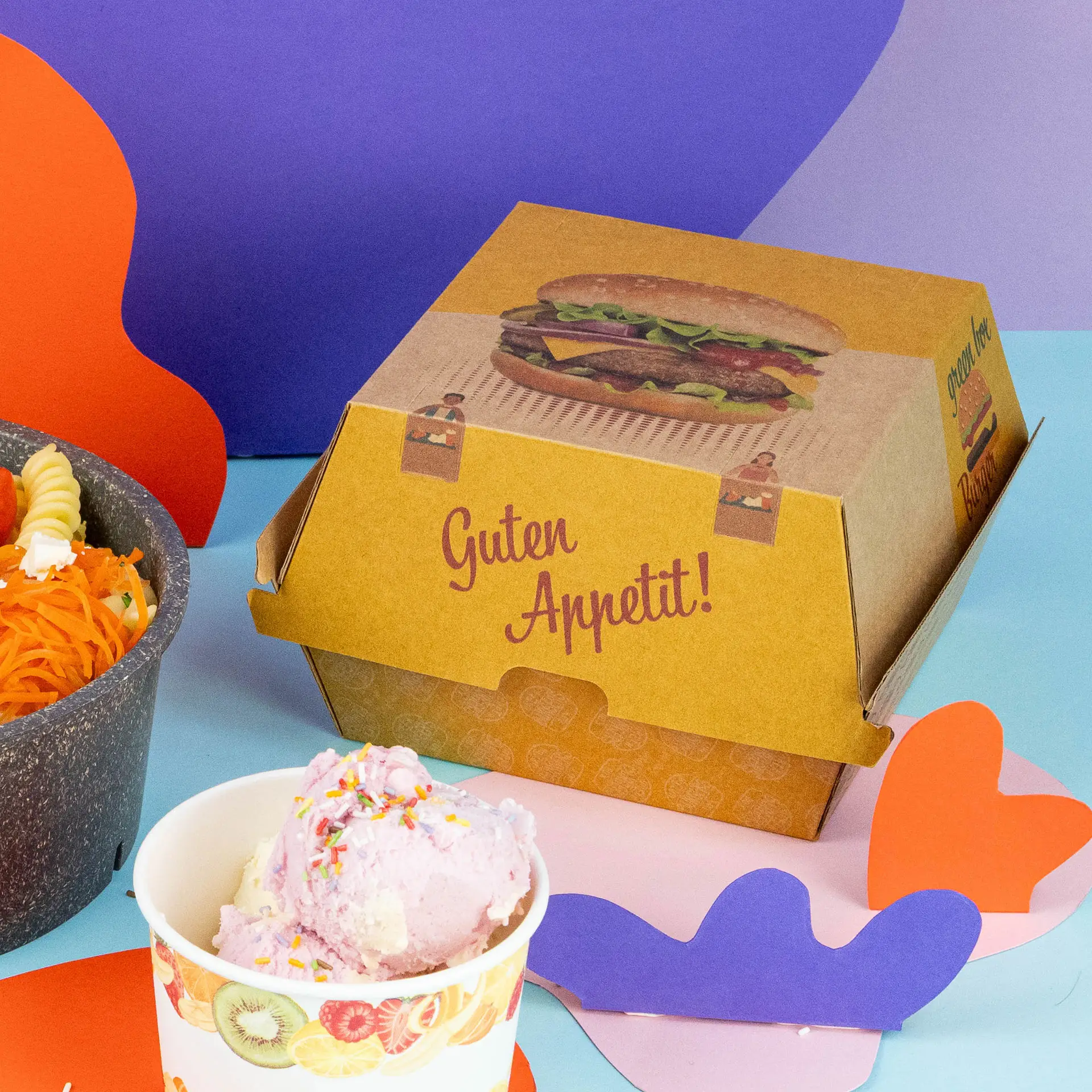 Scatola per hamburger marrone stampata con un grafico di hamburger, posizionata accanto a una ciotola di insalata e a un bicchiere di gelato rosa.